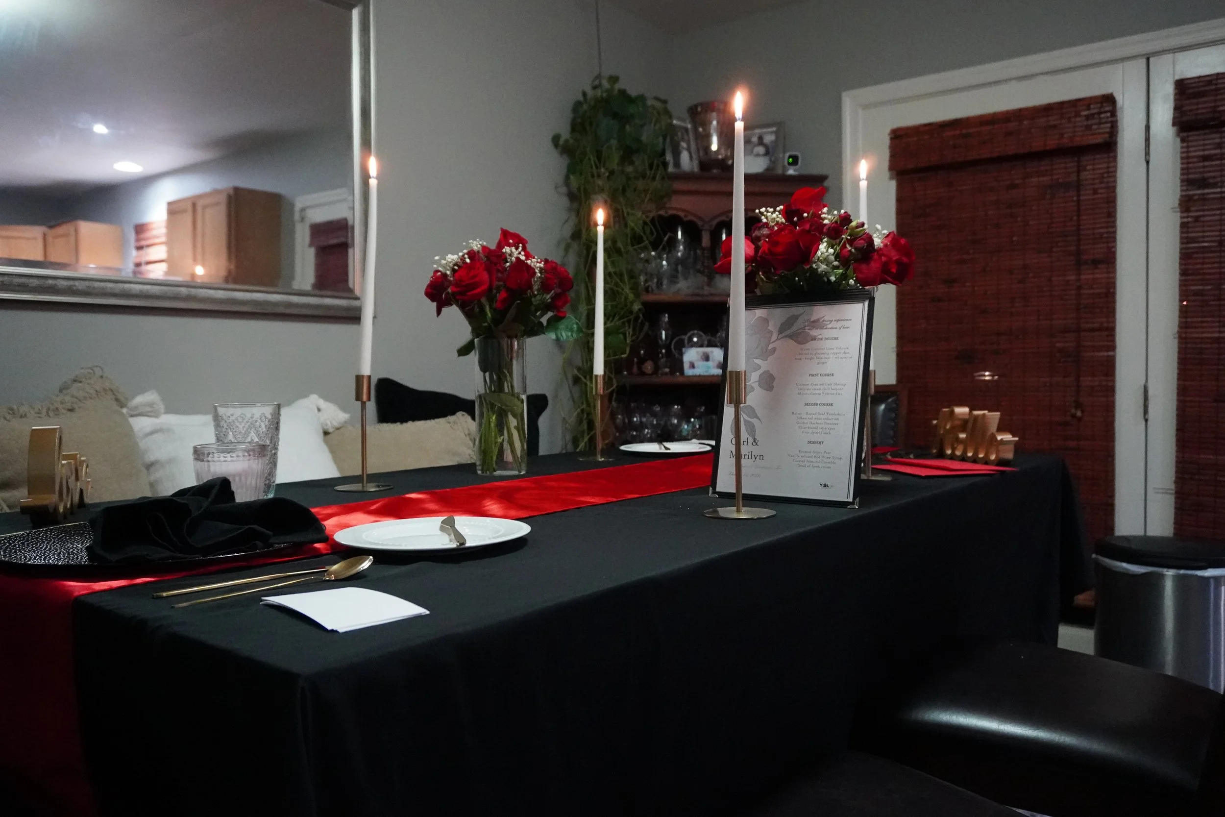 A dining table decorated with red roses in a vase, black tablecloth, and gold utensils, with candles and a menu frame, in a dimly lit room with a mirror and wooden blinds.