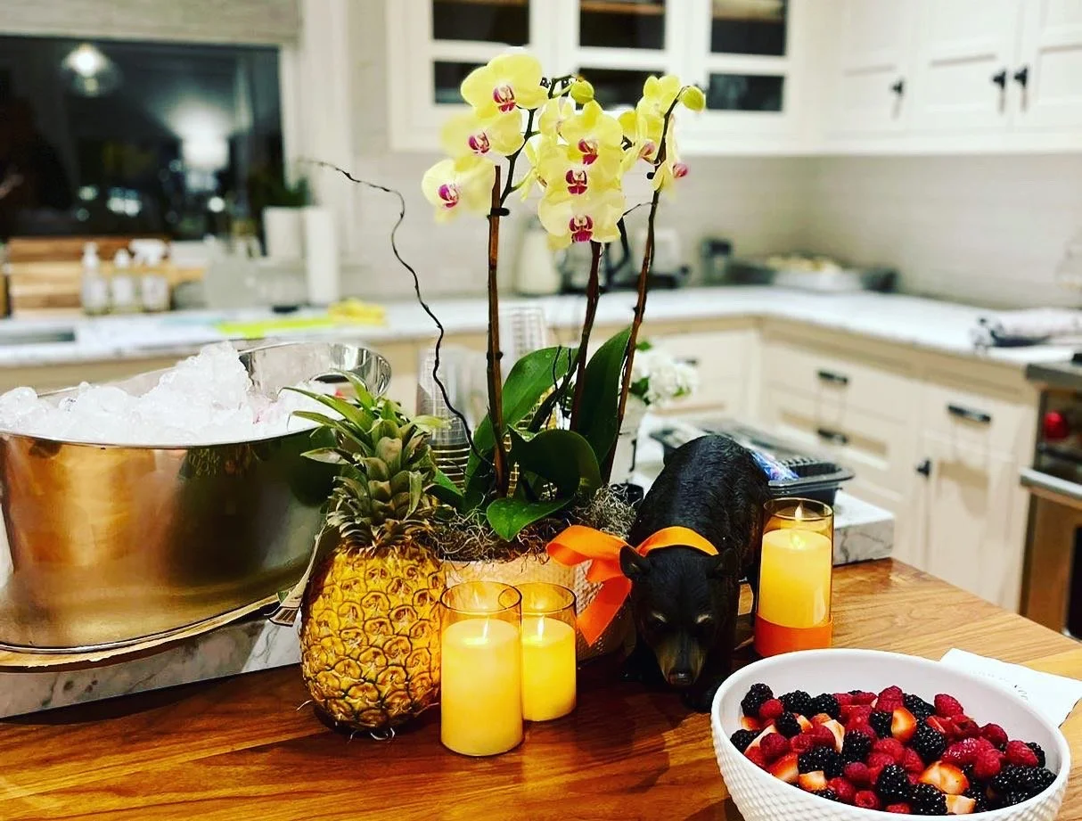 Decorative centerpiece with orchids, a pineapple-shaped container, and a black pig figurine with an orange ribbon, surrounded by candles, berries, and a bowl of mixed berries on a kitchen counter.