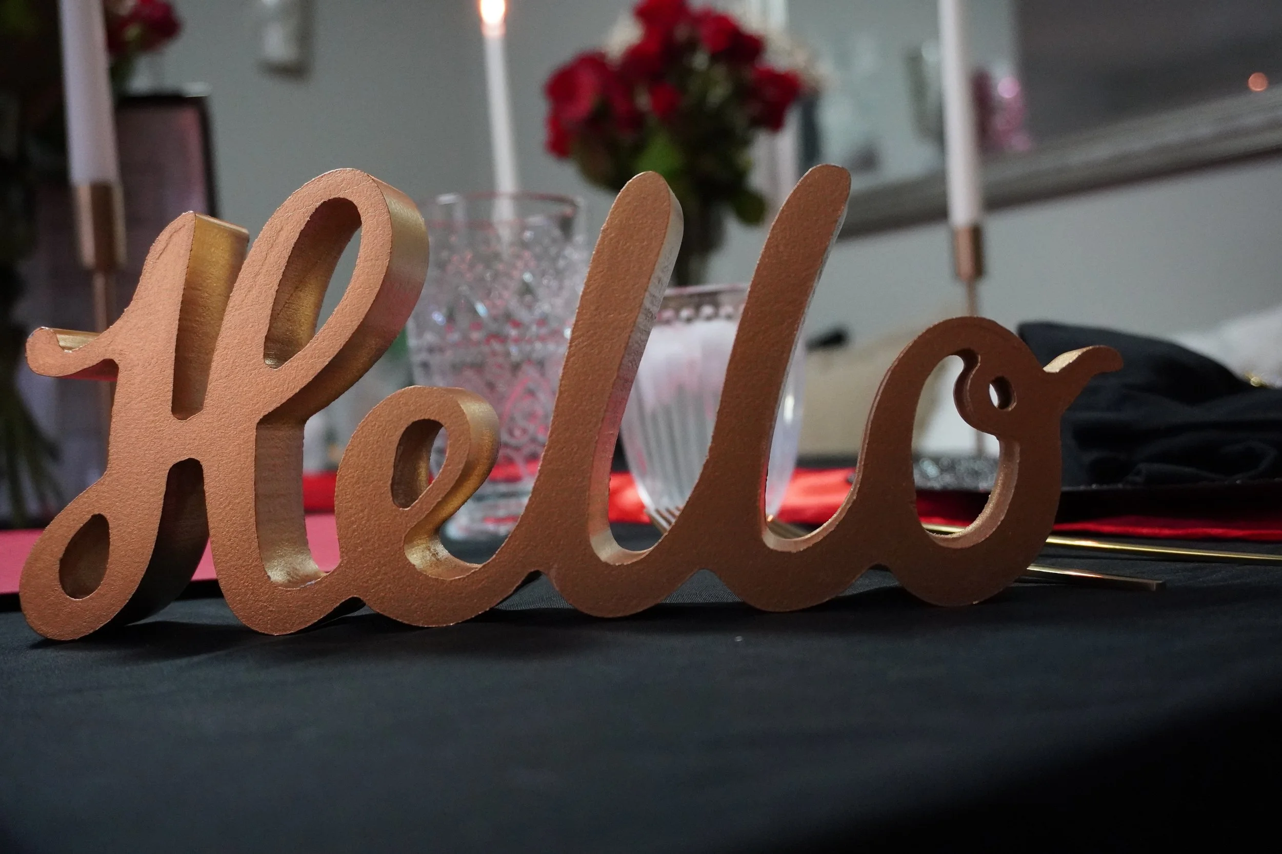 A brown wooden decoration spelling 'hello' placed on a black tablecloth with glasses, flowers, and candles in the background.