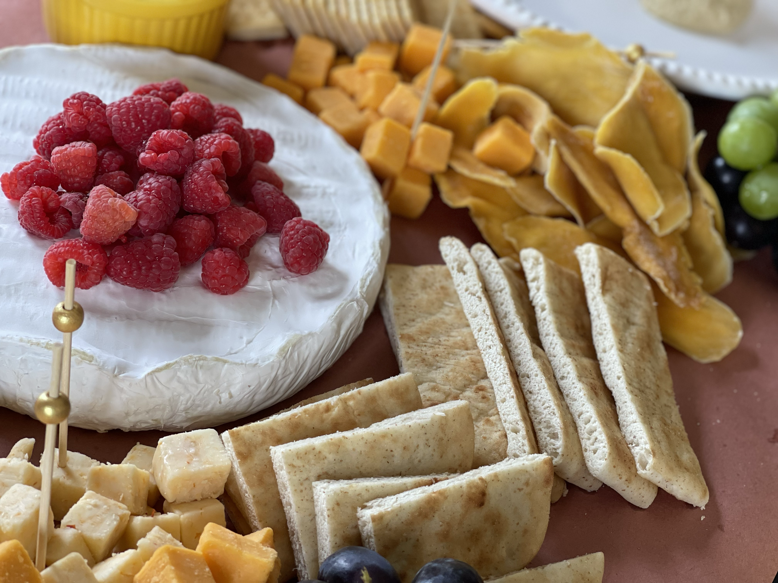 Cheese platter with brie topped with raspberries, sliced cheddar, crackers, grapes, dried mango, and a yellow dip.