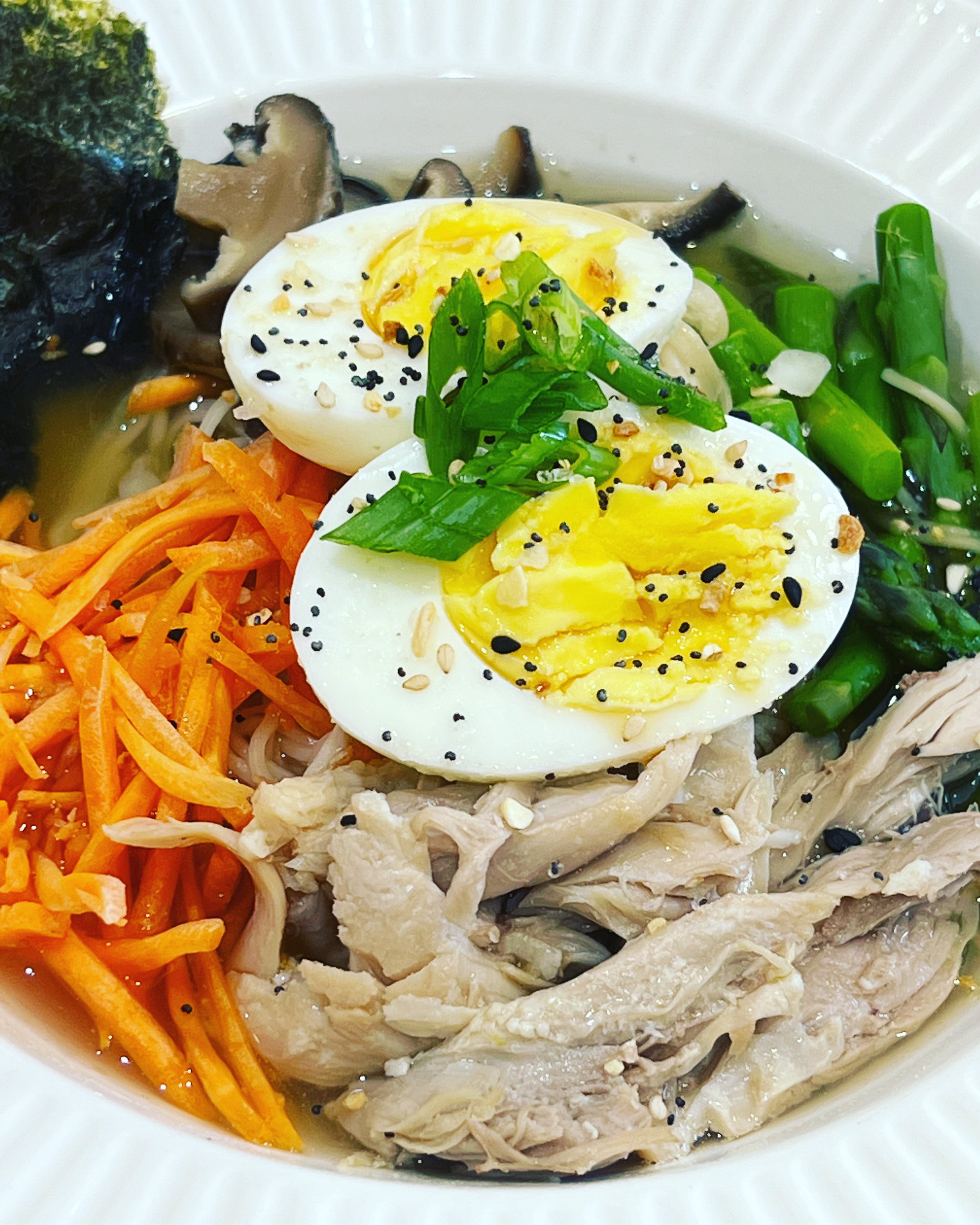 A bowl of Korean cold noodle soup with boiled eggs, shredded carrots, chicken, green onions, and sliced mushrooms, garnished with sesame seeds and black sesame seeds.