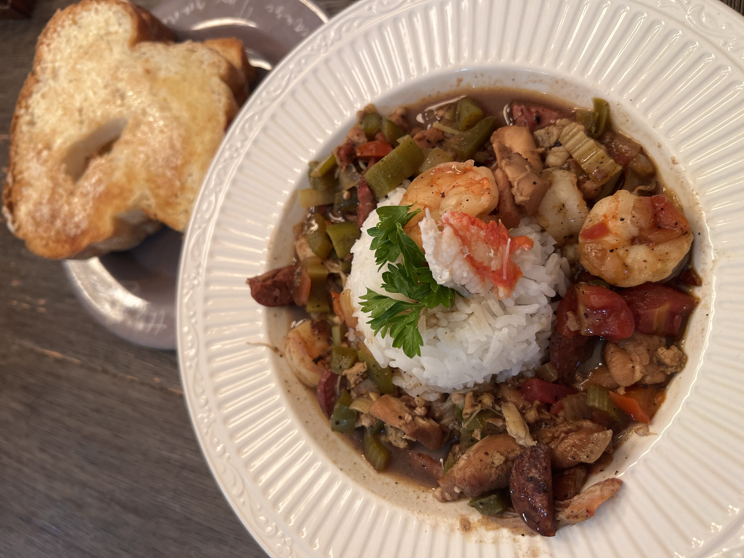A bowl of shrimp and vegetable stew with rice, garnished with parsley and a dollop of crab salad, served with toasted bread slices on a small plate.