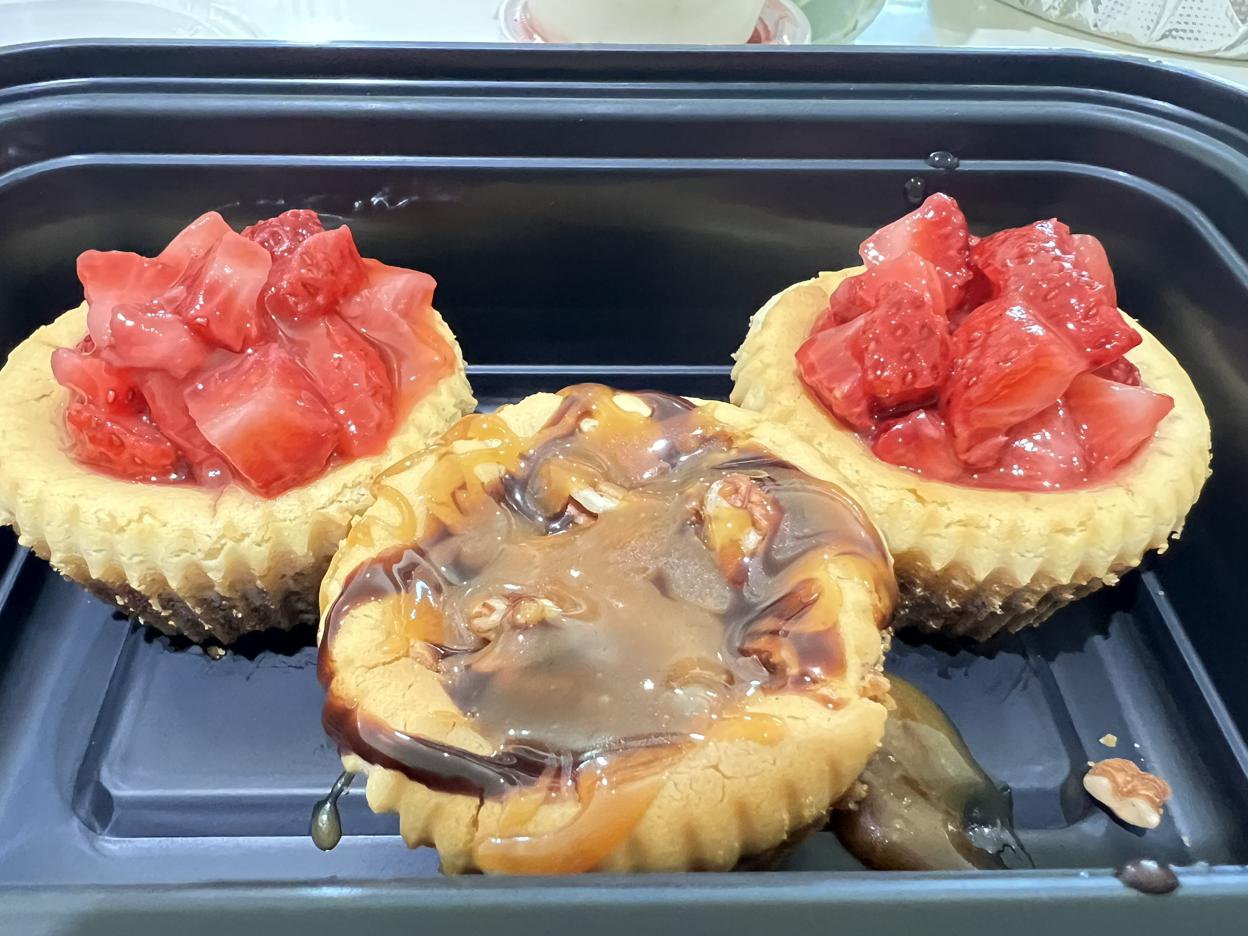 Three mini fruit tarts in a black plastic container. Two topped with chopped strawberries and one with caramel and chocolate drizzle.
