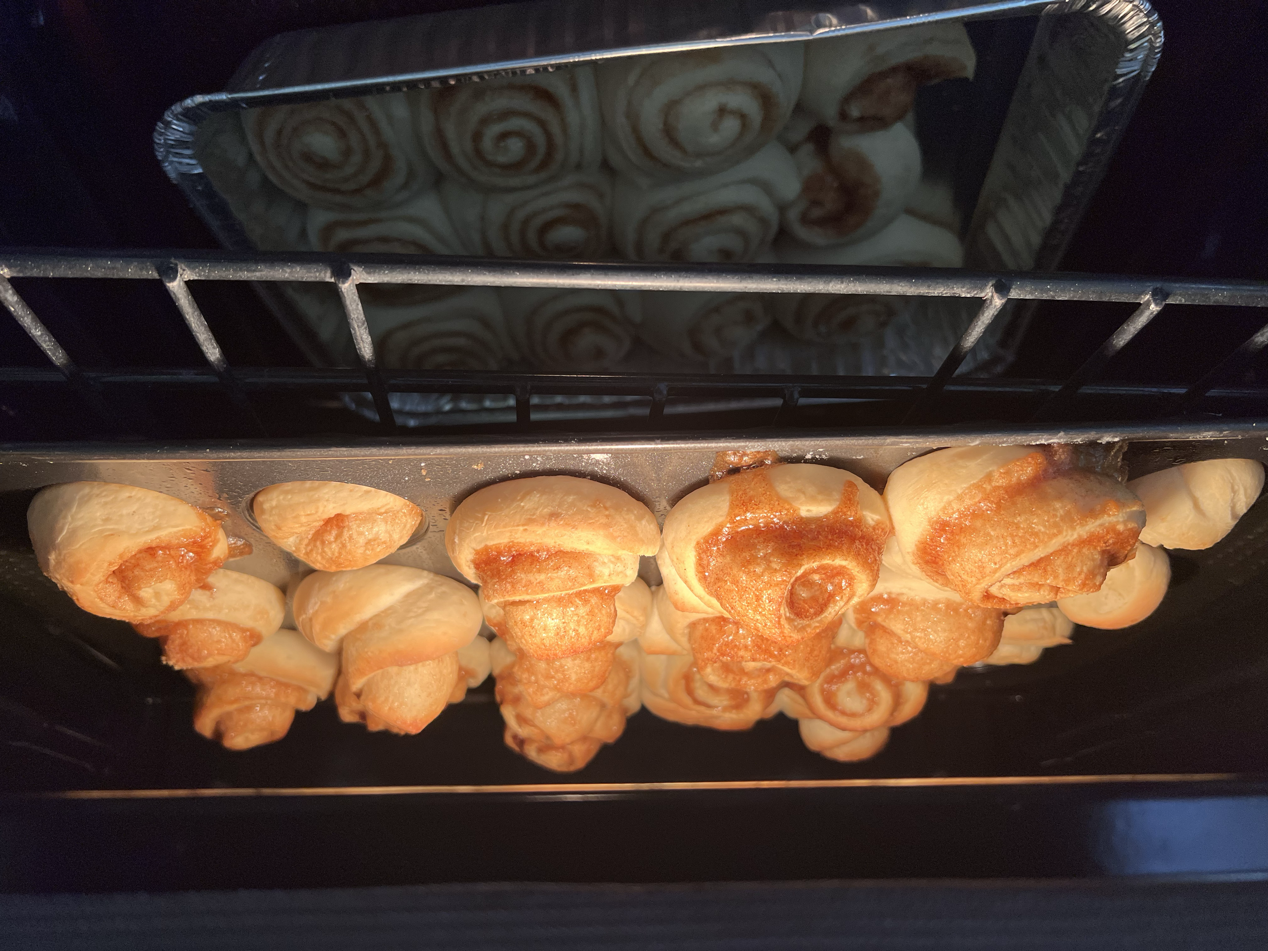 Oven with cinnamon rolls baking on the bottom rack and more cinnamon rolls cooling on a tray on the top rack.