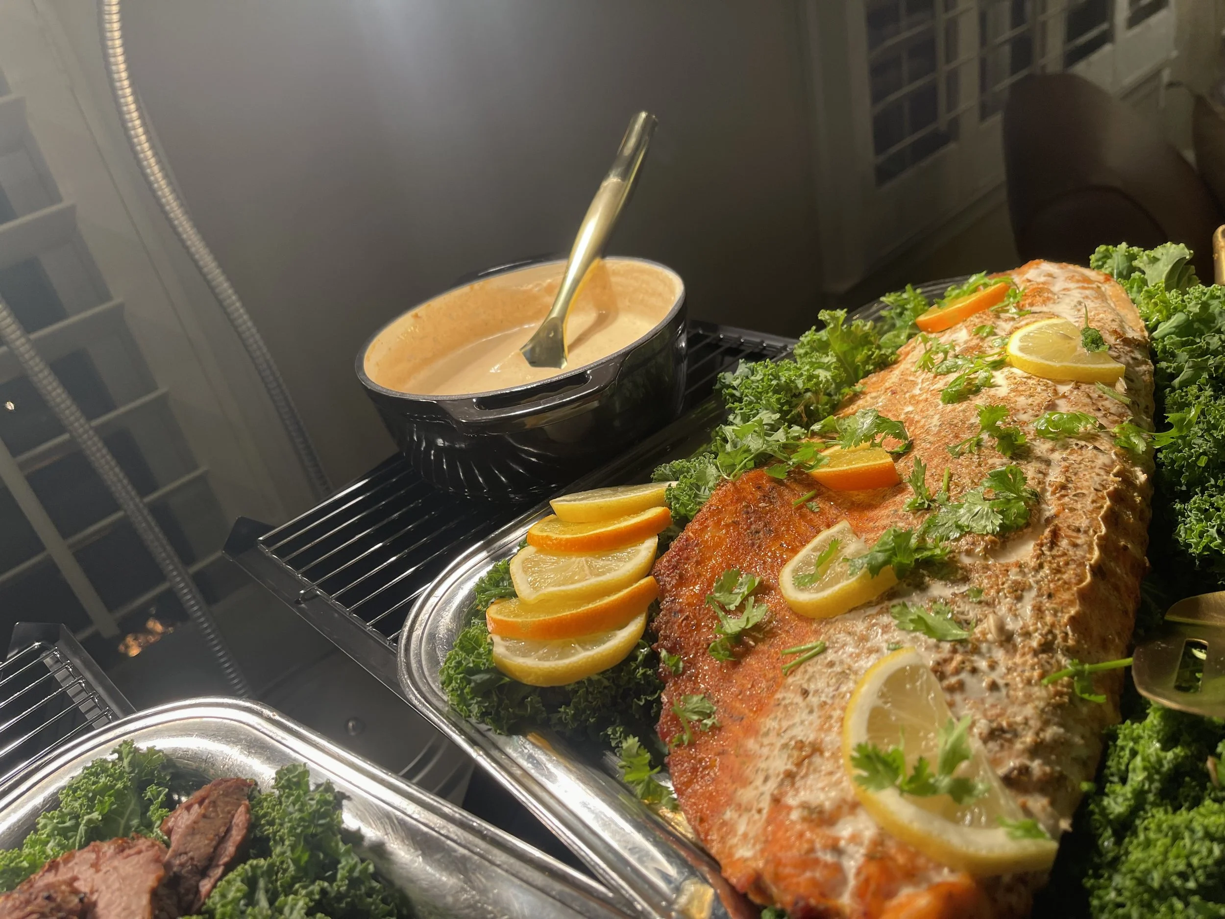Baked salmon garnished with lemon slices, parsley, and vegetables, with a bowl of sauce nearby on a buffet table.
