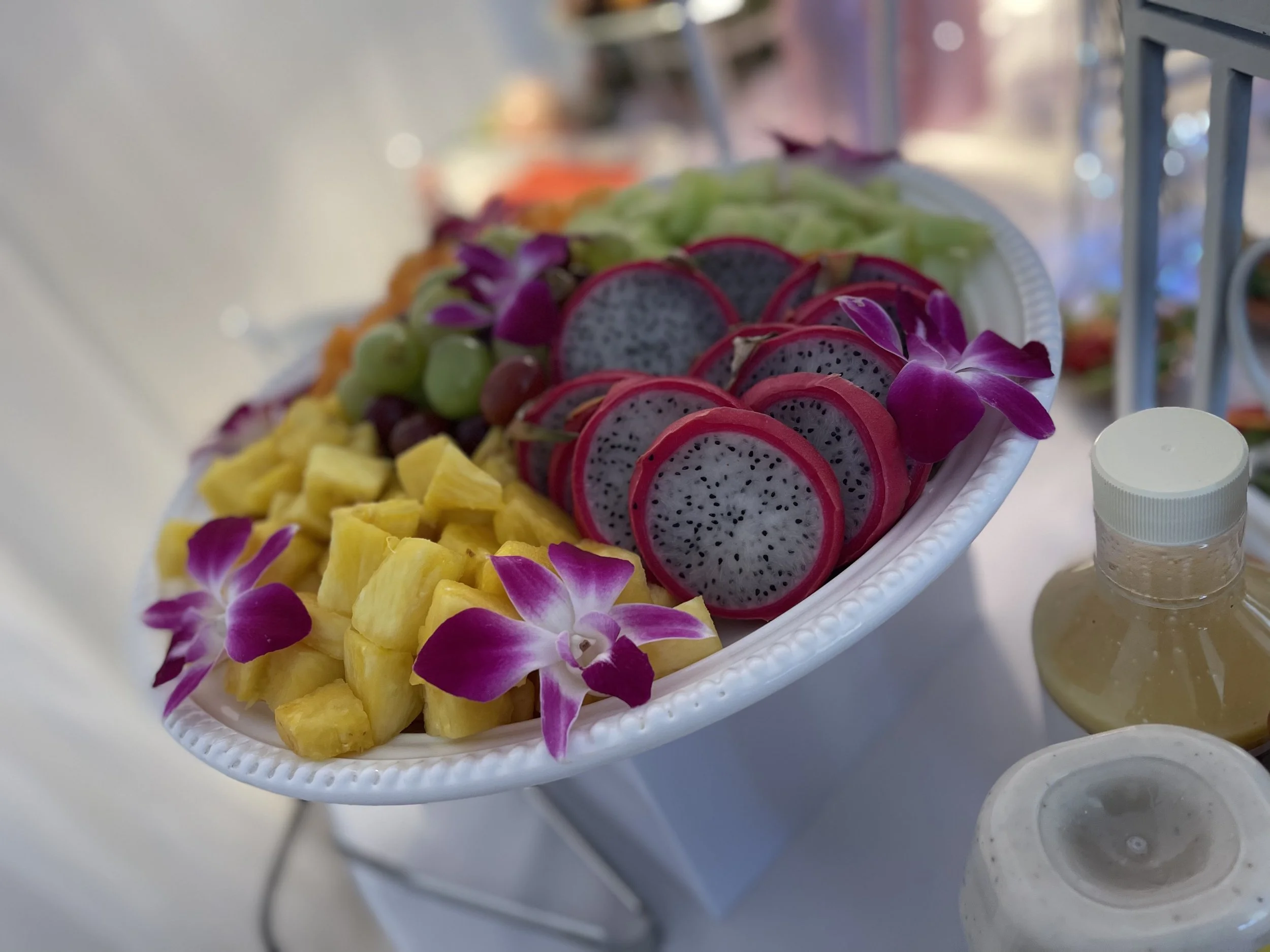 A platter of assorted fresh fruit including pineapple chunks, sliced dragon fruit, green and red grapes, and lettuce, decorated with purple orchids.