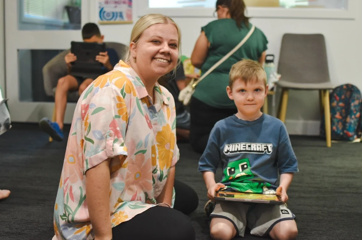 A woman and a young boy posing for a picture in a room with other people, some sitting and using tablets, and backpacks nearby.