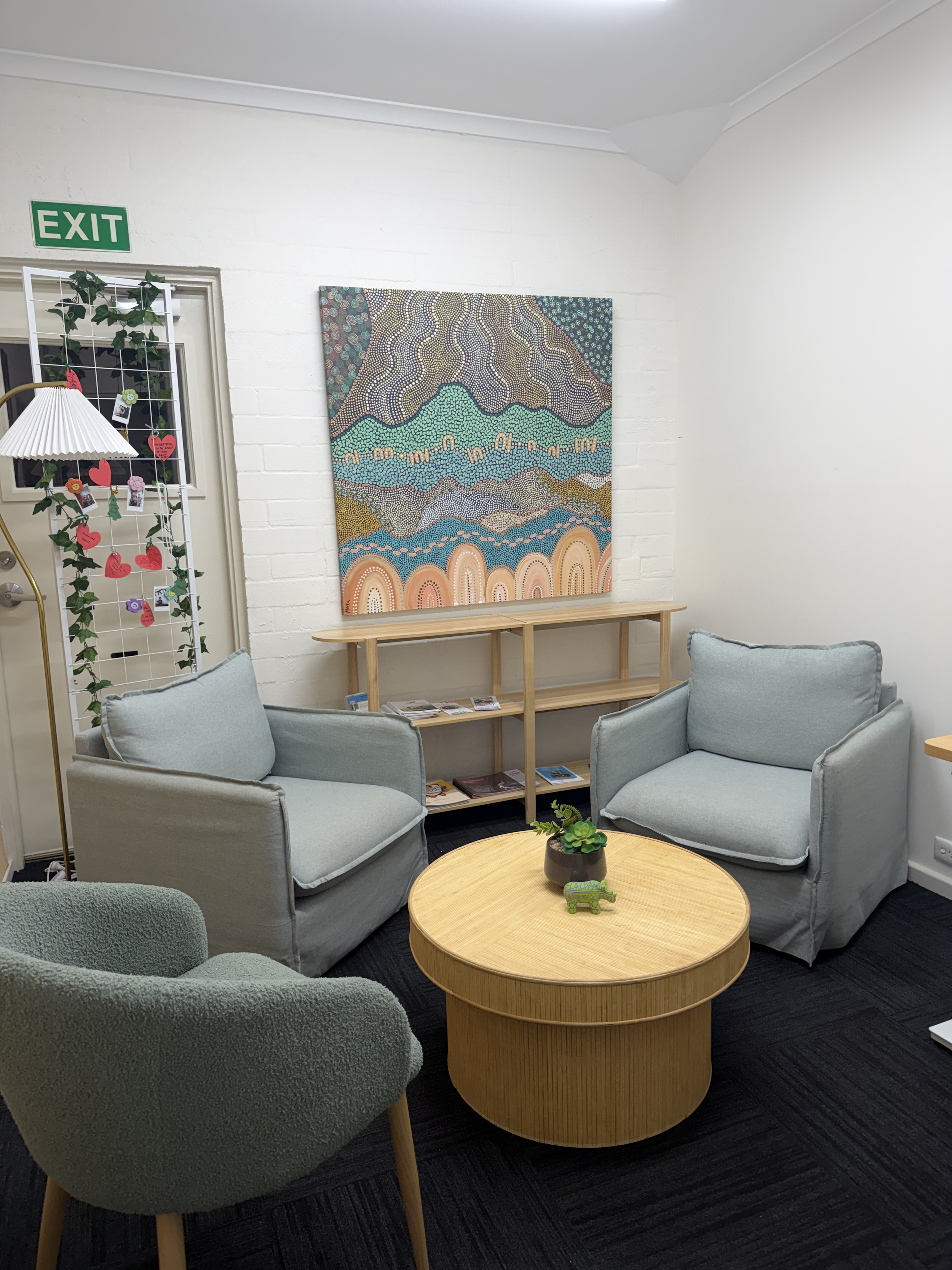 A cozy sitting area with two light gray armchairs, a dark green accent chair, and a round wooden coffee table decorated with small plants and a decorative toy. A colorful abstract painting hangs on the white brick wall behind the chairs, and beside the door with an 'EXIT' sign, there is a decorative wooden rack with hanging red paper hearts and photos.