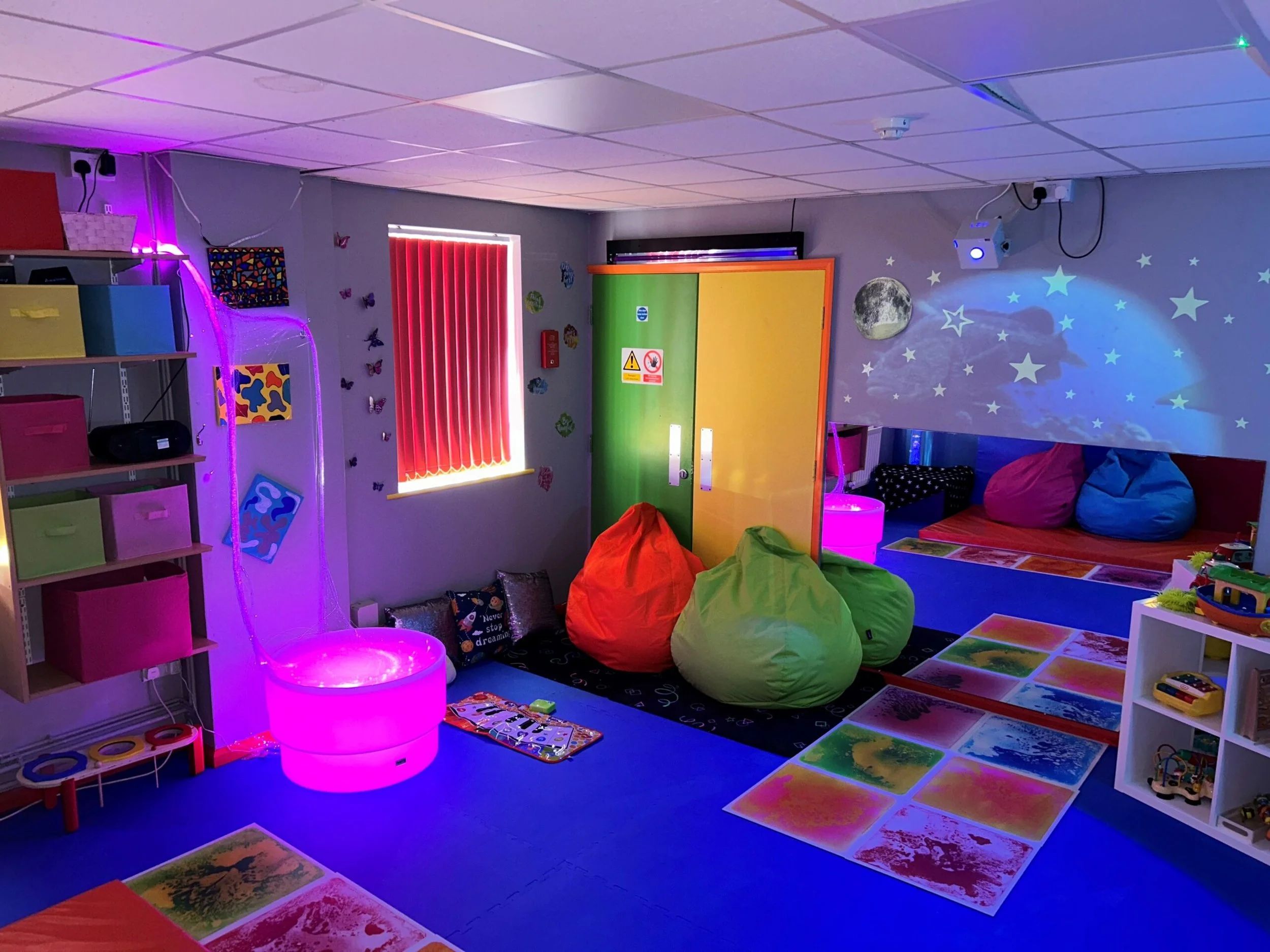 Colorful children's playroom with glow-in-the-dark star and moon decorations, bean bag chairs, soft mats, and ambient purple and pink lighting.