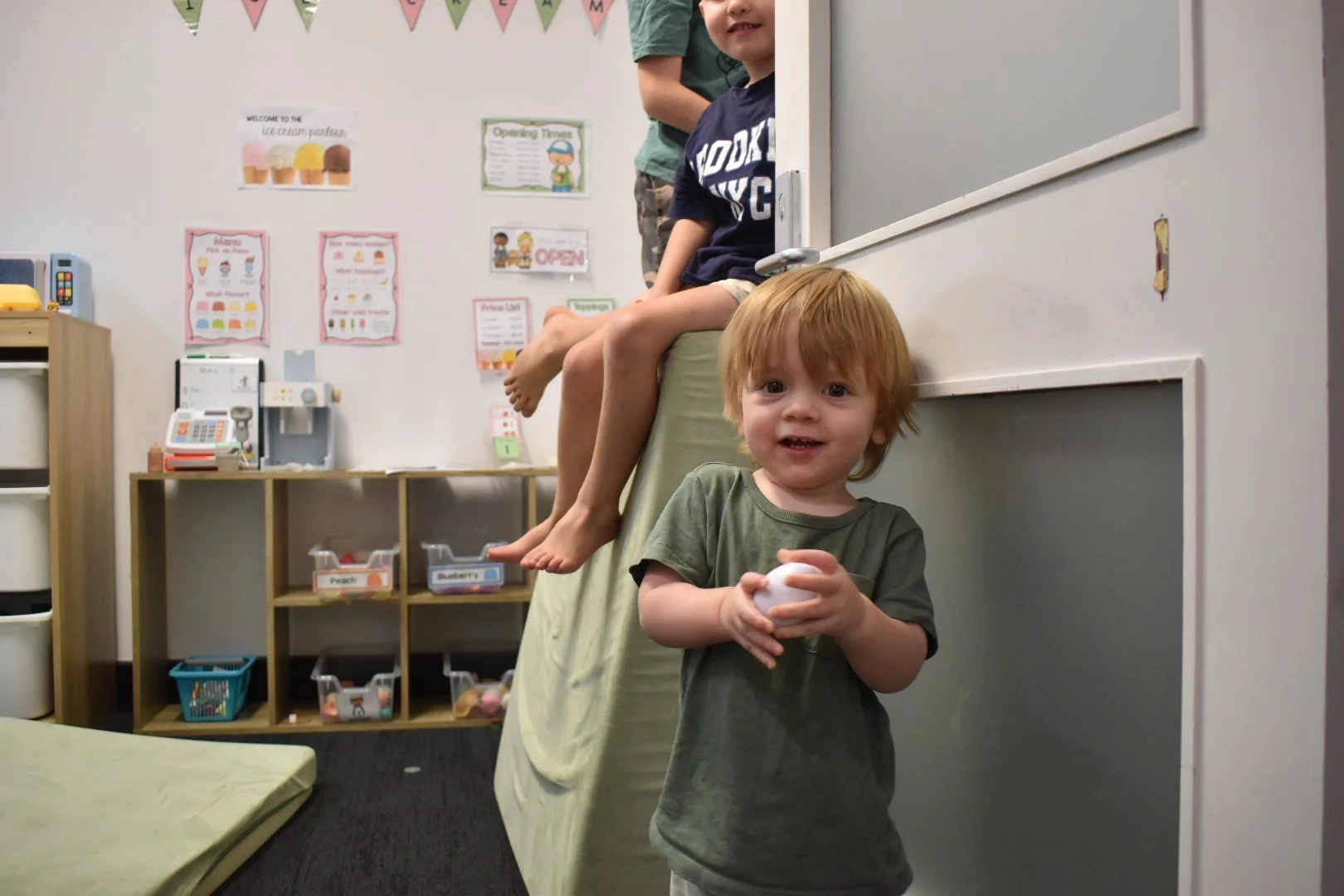 A young child with red hair smiles while holding a small white ball in a classroom. Behind him, two other children are sitting on a cushioned ledge, with a classroom wall decorated with colorful posters, including a menu and opening times, and cubbie