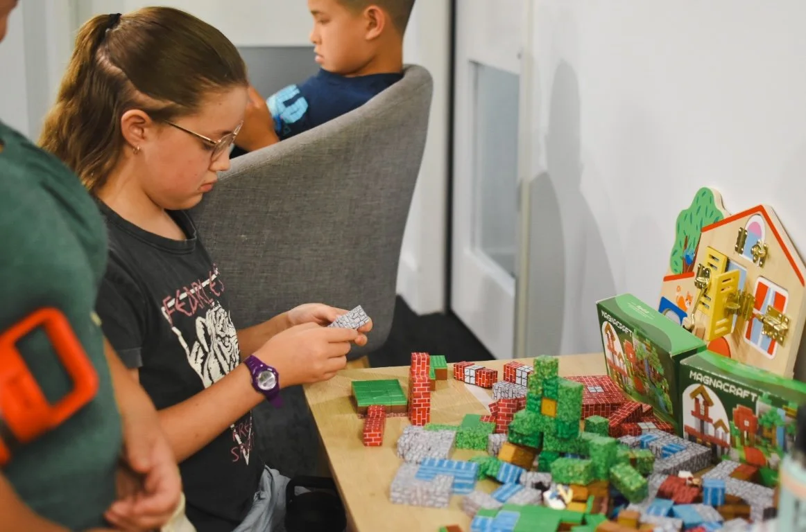 A girl with glasses and a purple watch plays with colorful building blocks while a boy in a green shirt observes. There are various toys and games on the table.