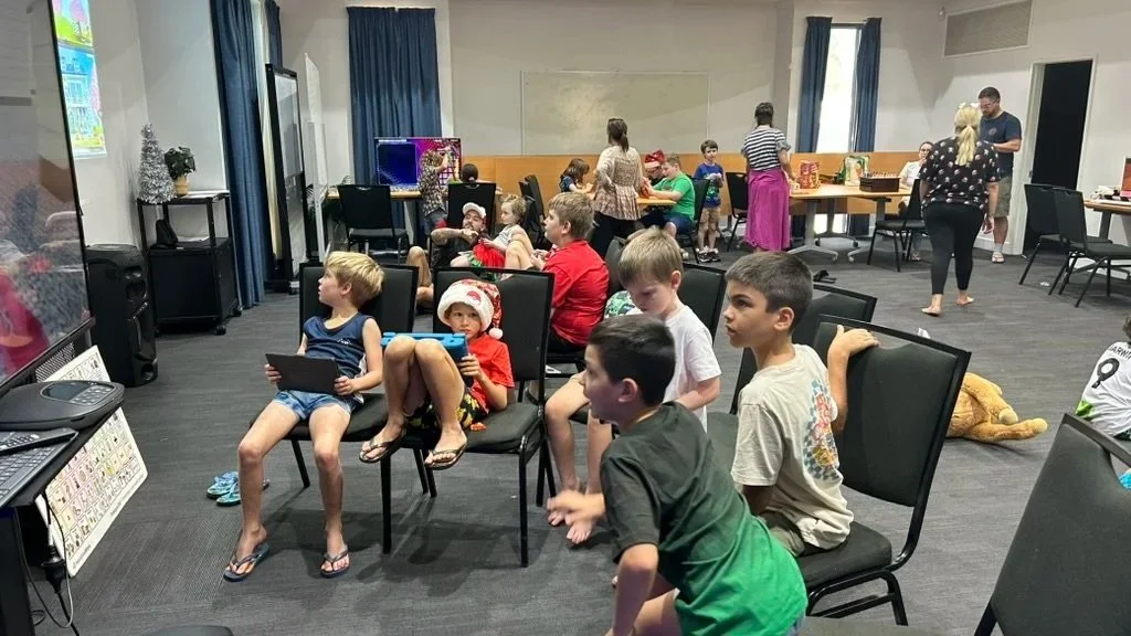 Children sitting in chairs watching a screen, with Christmas decorations and adults in the background, indicating a holiday party or gathering.