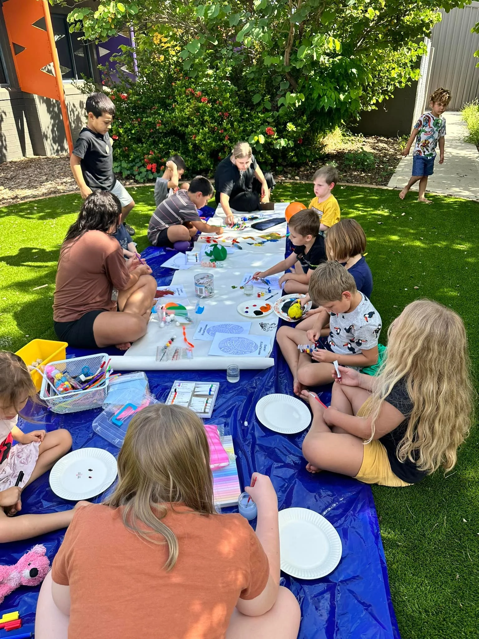 Children and adults sitting on a blue tablecloth in a backyard, engaging in arts and crafts activities with various supplies. Some children are drawing, while adults assist. The setting is sunny with green grass, trees, and bushes in the background.