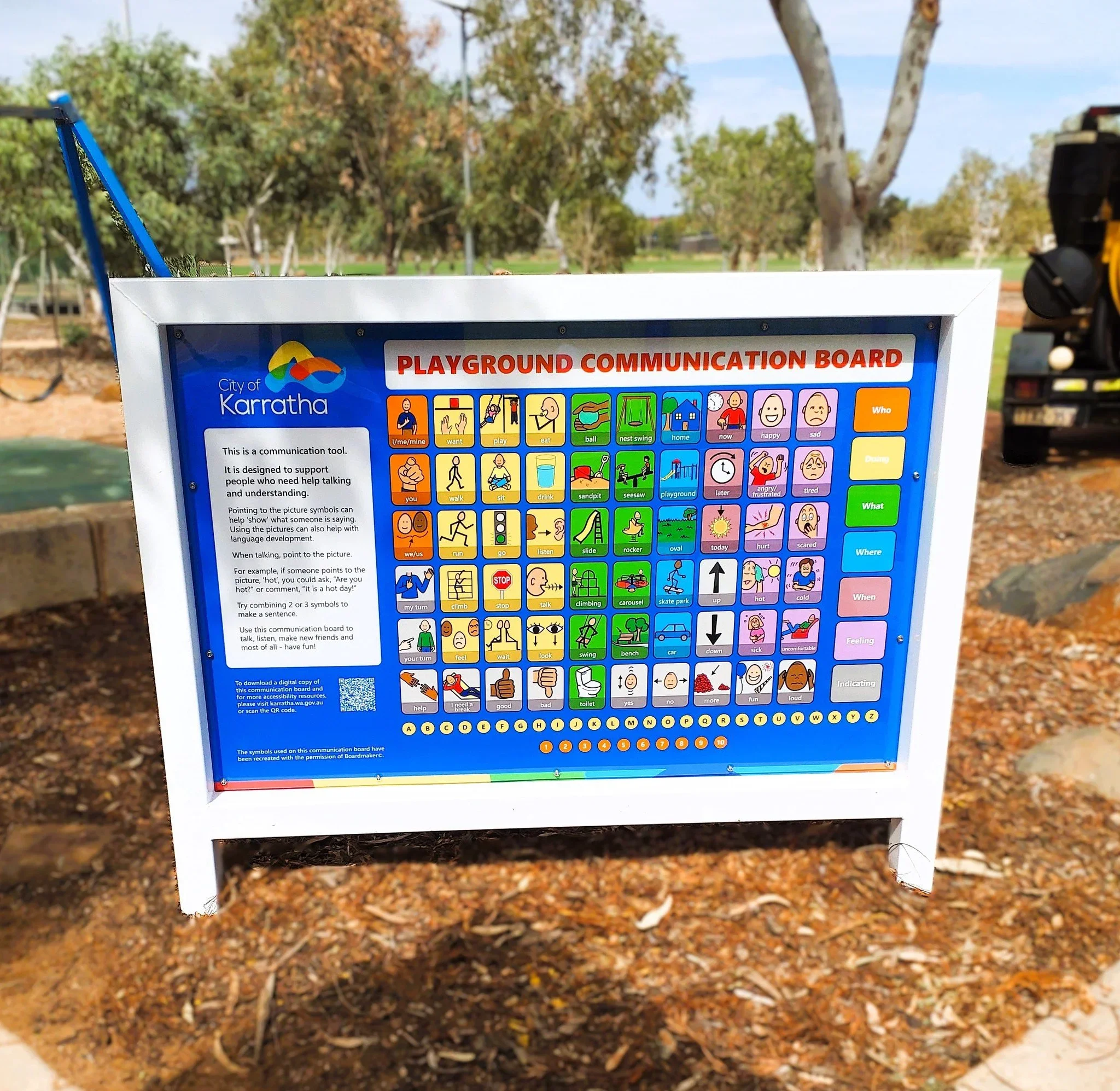 A colorful playground communication board with pictures and symbols for children to express feelings, actions, and needs, located outdoors with trees and a clear sky in the background.