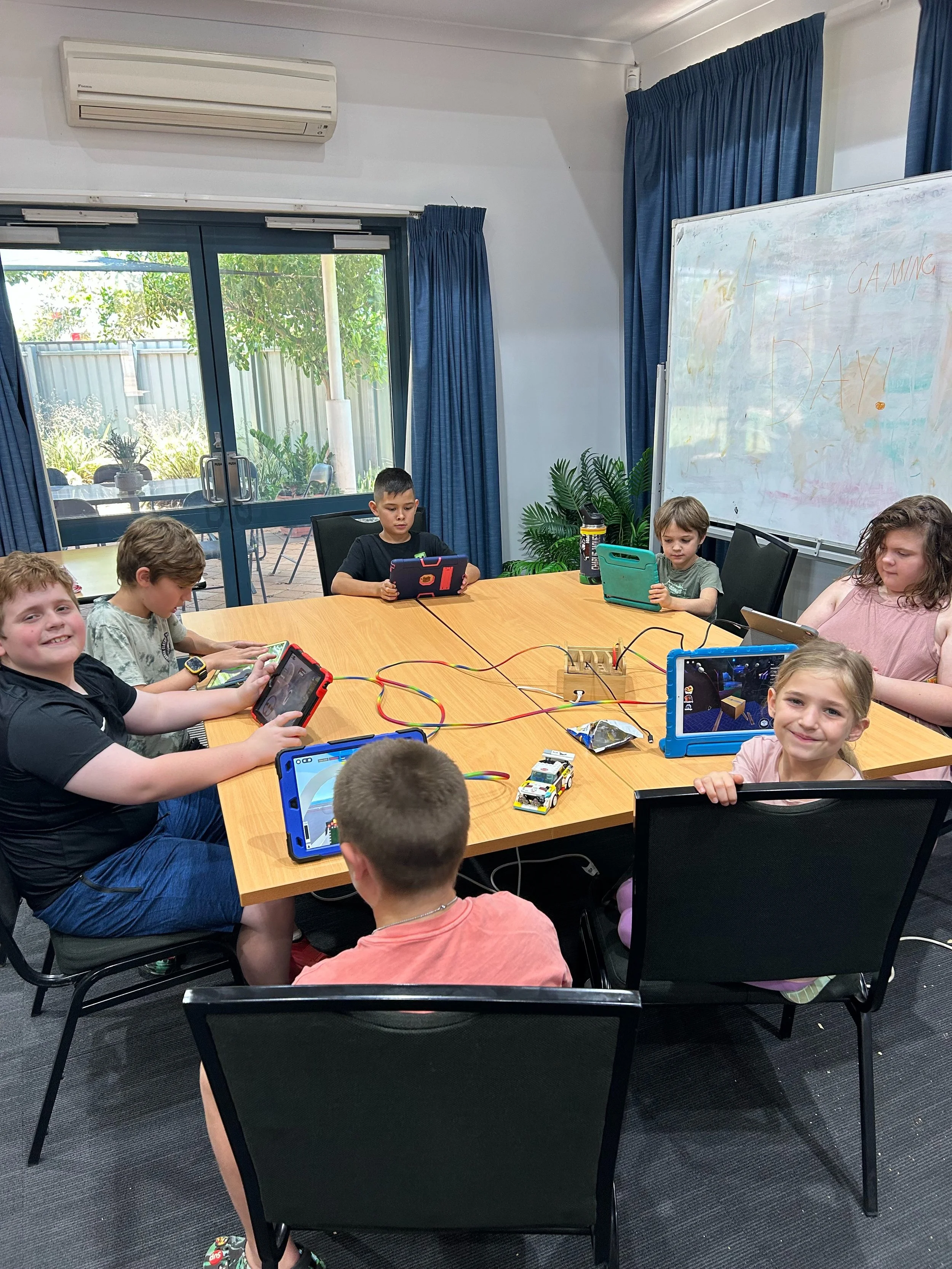 Children sitting around a rectangular table engaged in educational activities using tablets and robotics kits in a classroom with a whiteboard and large sliding glass doors leading outside.