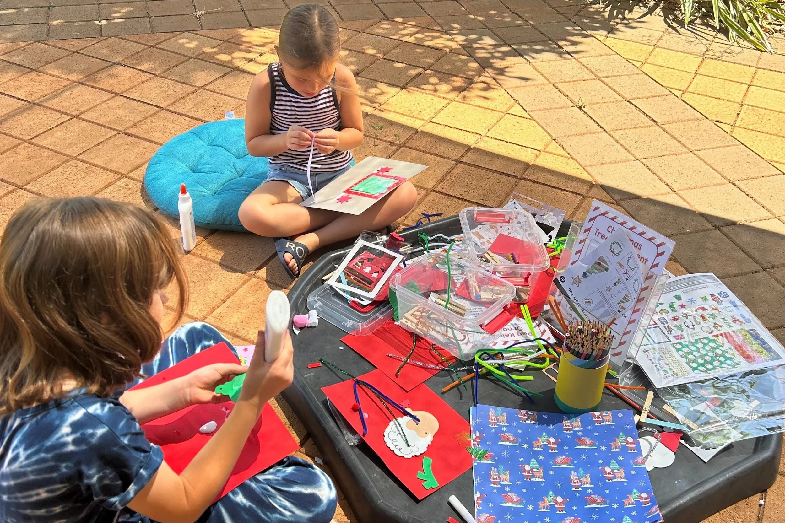 Two young girls sitting outdoors on paving bricks, engaged in Christmas craft activities, with craft supplies spread on a table and the girl in the background drawing on paper.