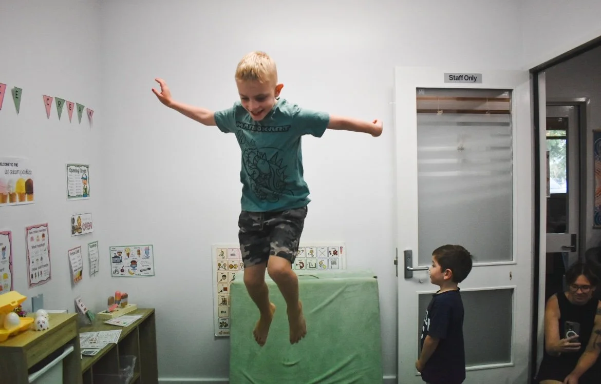 A young boy is jumping in a room, smiling with arms outstretched, while two other children and an adult watch.