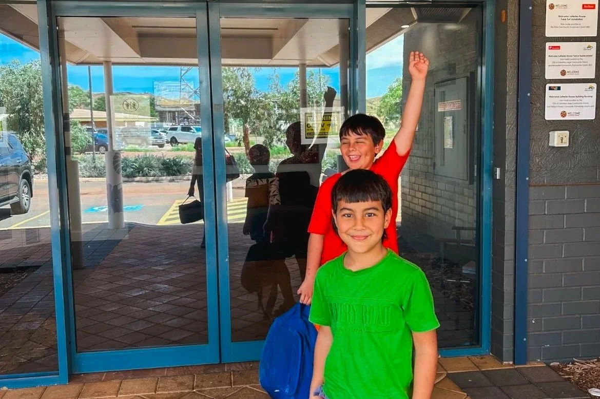 Two boys standing outside a glass door, smiling. One in a red shirt has his arm raised, the other in a green shirt holds a blue bag. Reflection of people and cars can be seen in the glass.