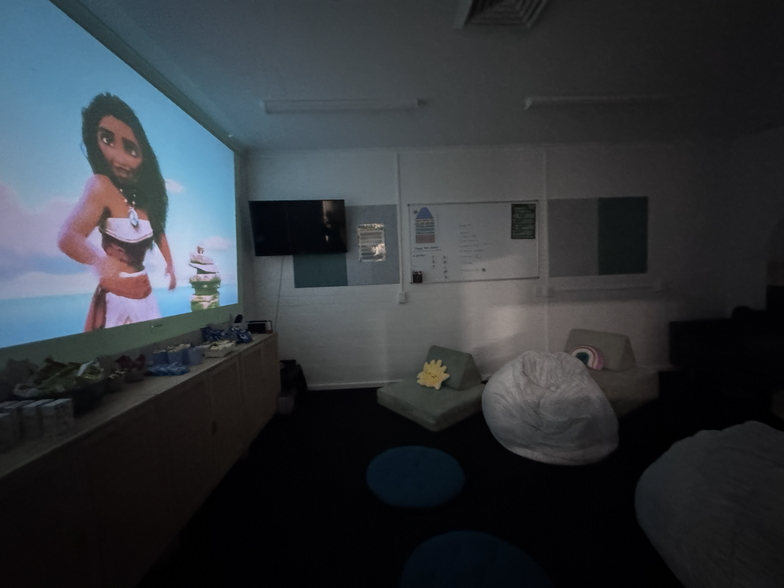 A dark room with a large screen projecting an animated image of a woman with dark, curly hair, wearing a white top and a necklace, standing outdoors with a blue sky and clouds in the background.