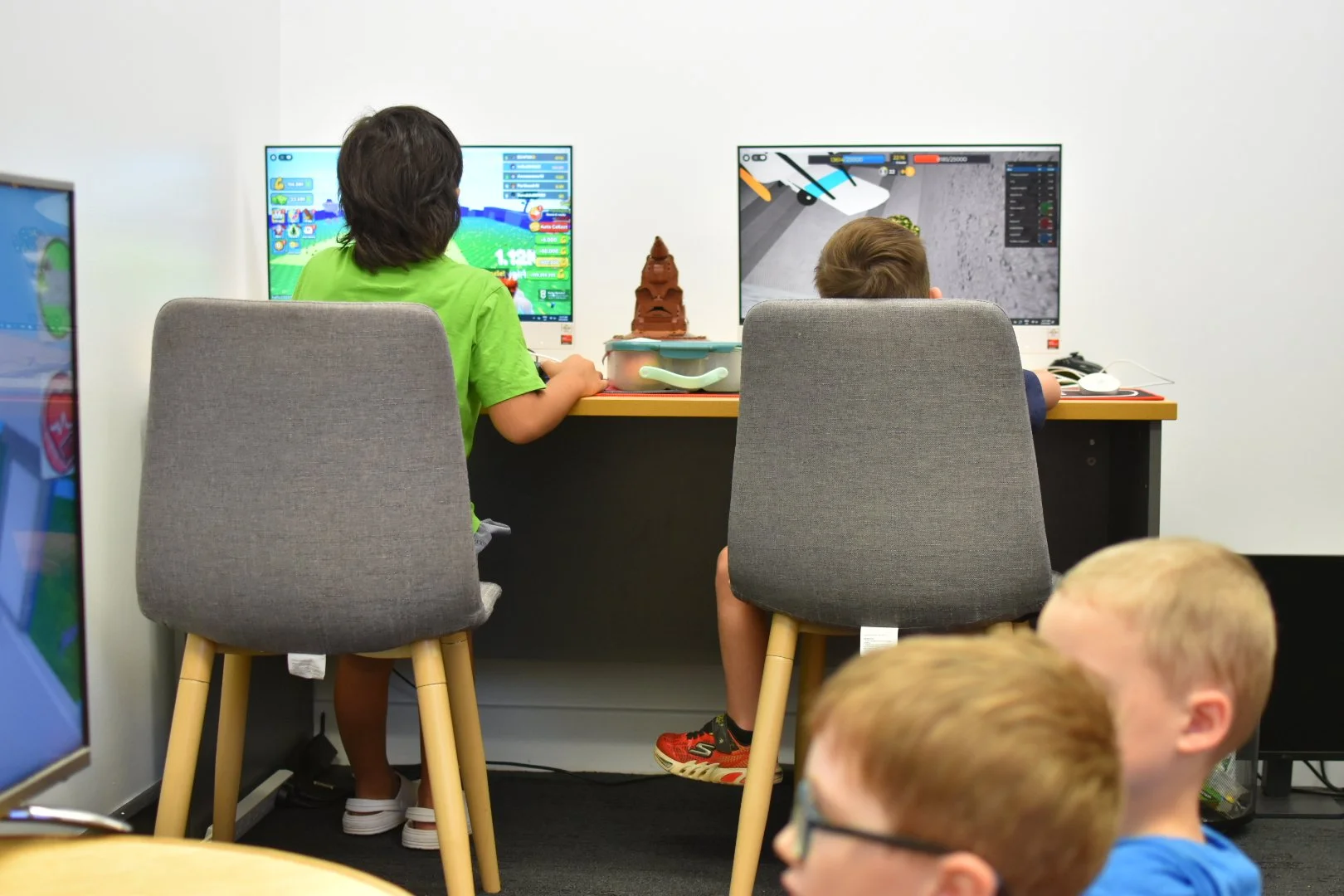 Two children sitting at a desk playing video games on computer monitors, with other kids in the foreground.