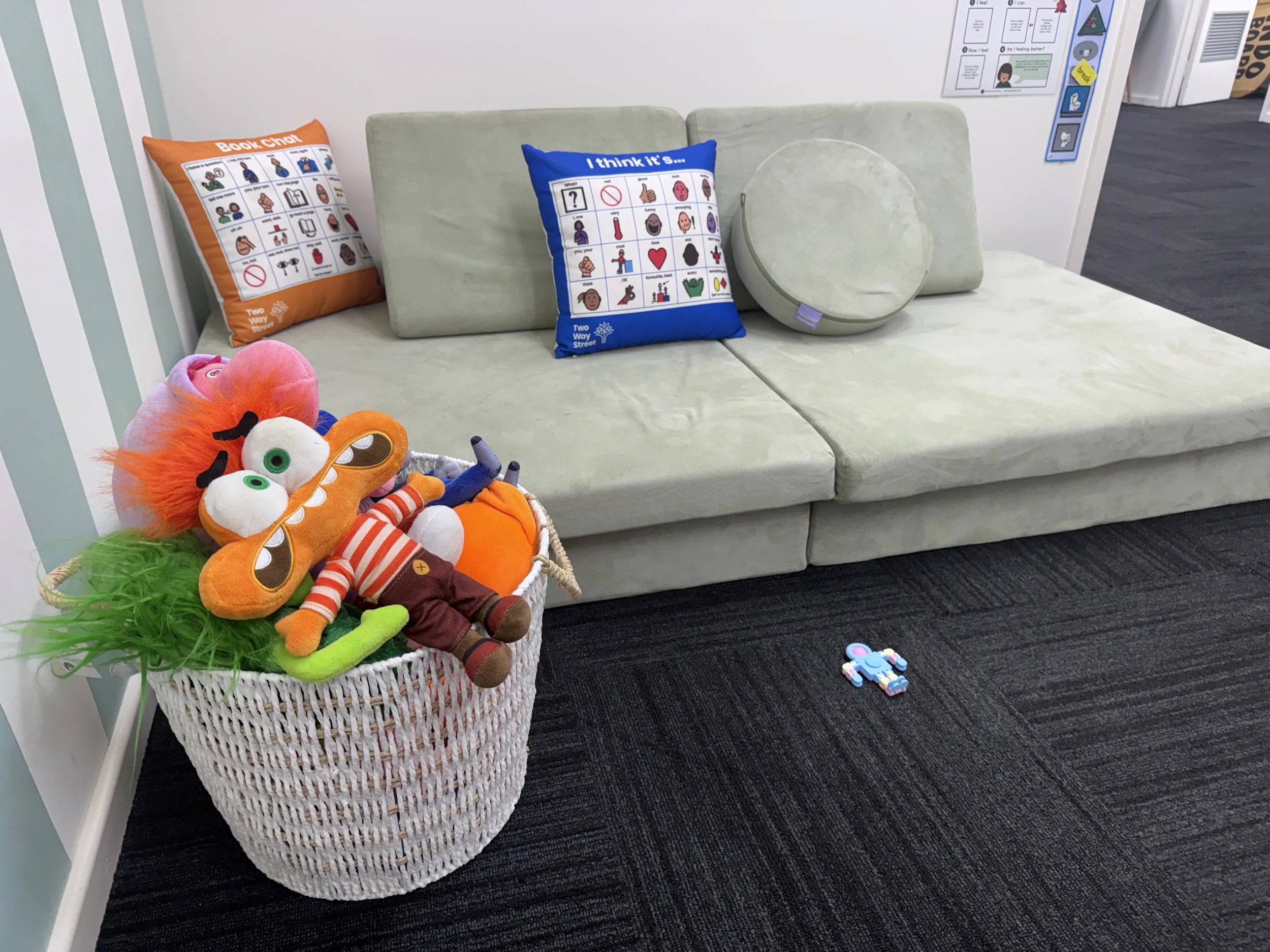 A waiting area with a light-colored couch, decorative pillows, a basket of plush toys, a puzzle piece on the dark carpet, and informational posters on the wall.