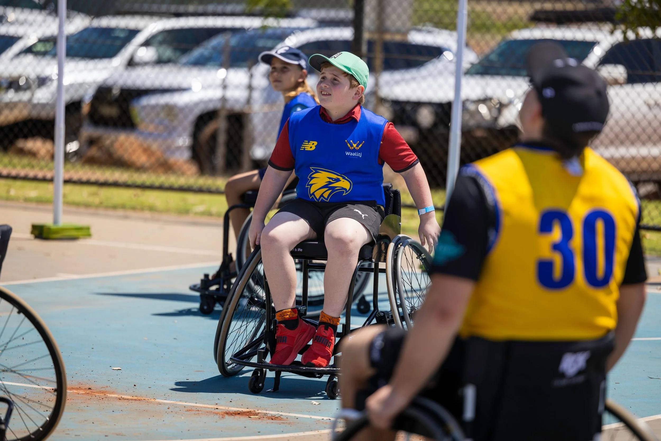 Children participating in a wheelchair basketball game outdoors, with a child in a wheelchair wearing a blue jersey and two children outside the wheelchair wearing yellow and black jerseys. A chain-link fence and parked cars are in the background.