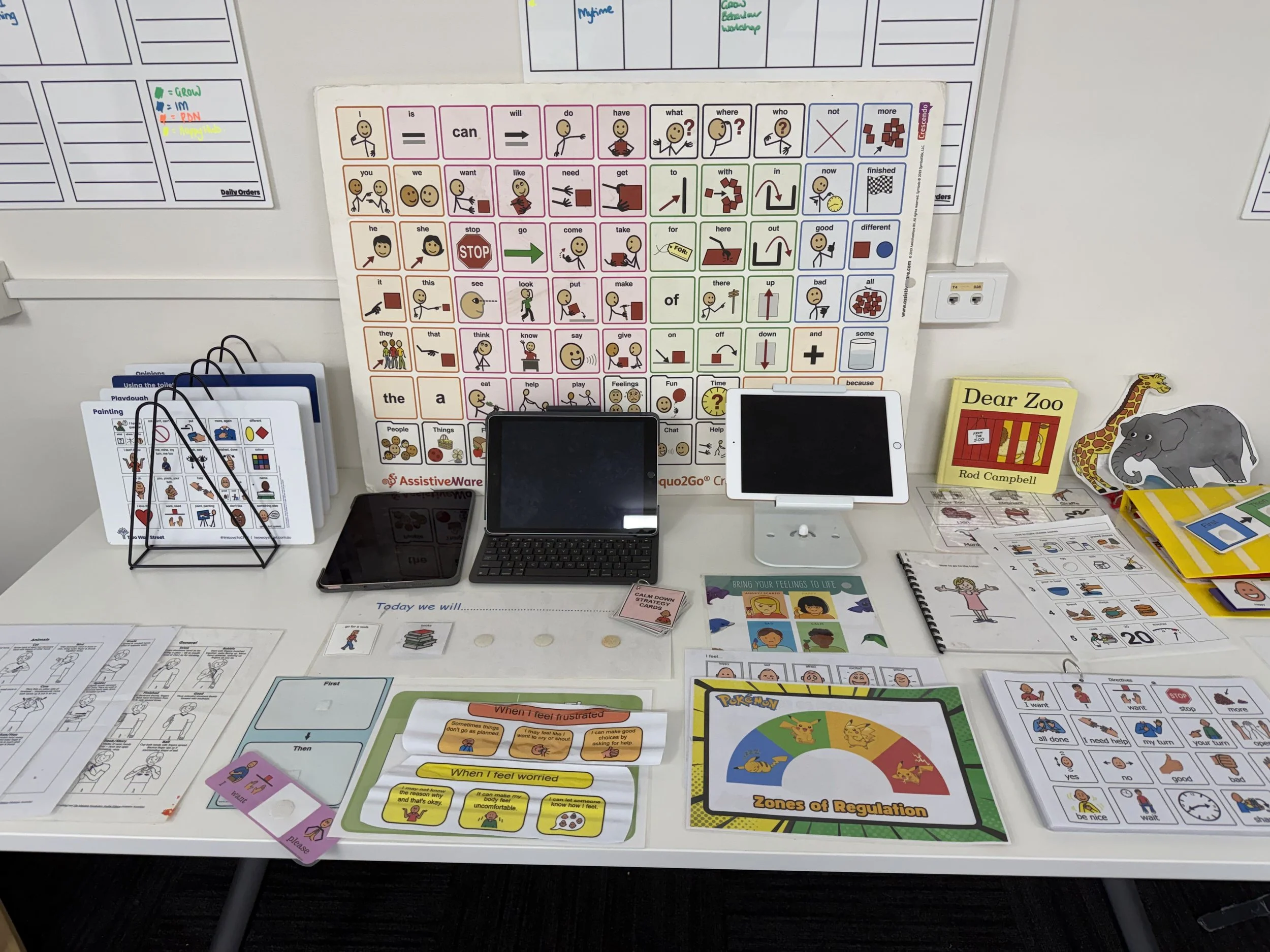 A desk with various educational materials, including communication boards, books, electronic tablets, and visual aids for learning and communication, likely in an educational or therapy setting.