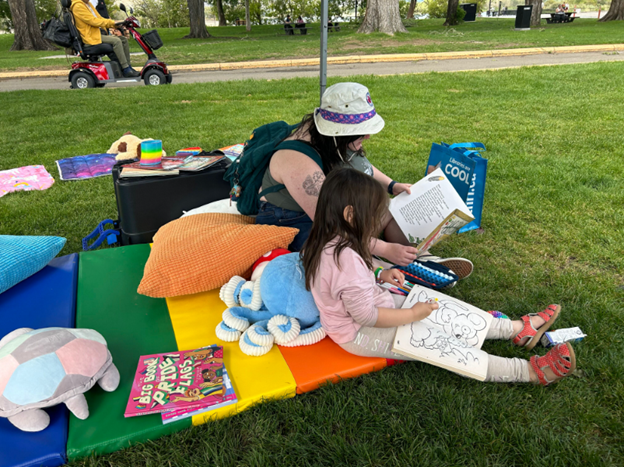 A woman and young girl sitting on a colorful outdoor mat in a park. The woman is reading a book while the girl is drawing on a sketchpad. Surrounding them are pillows, toys, and books. In the background, there is a person riding a mobility scooter and a park with trees.