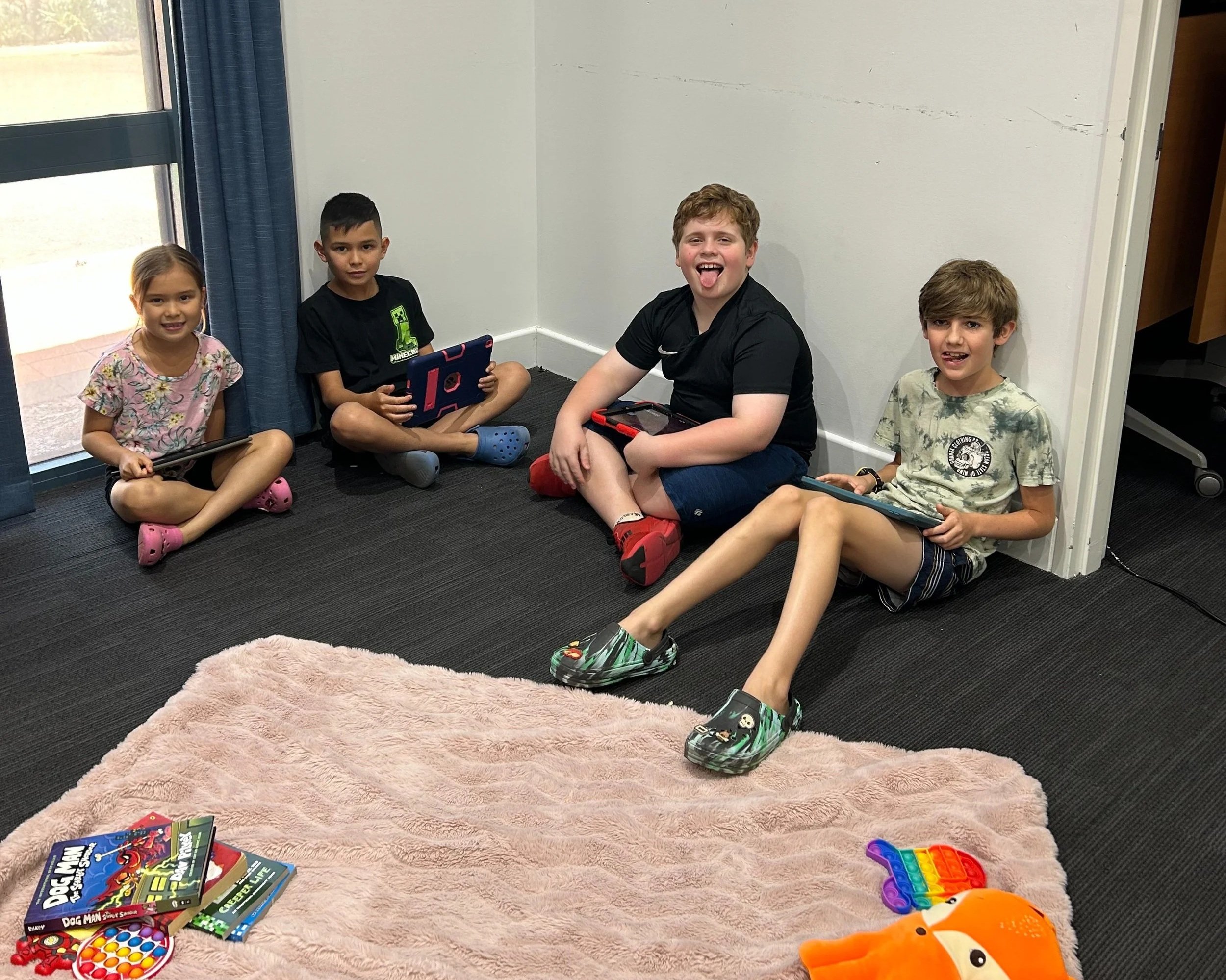 Five children sitting on the floor in a corner of a room, with some toys and books in front of them. They are posing for the photo, some smiling and some making faces.
