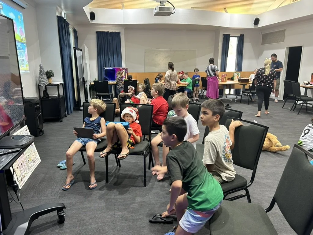 Children sitting and watching a large screen in a room decorated for Christmas, with adults in the background preparing snacks or playing games.