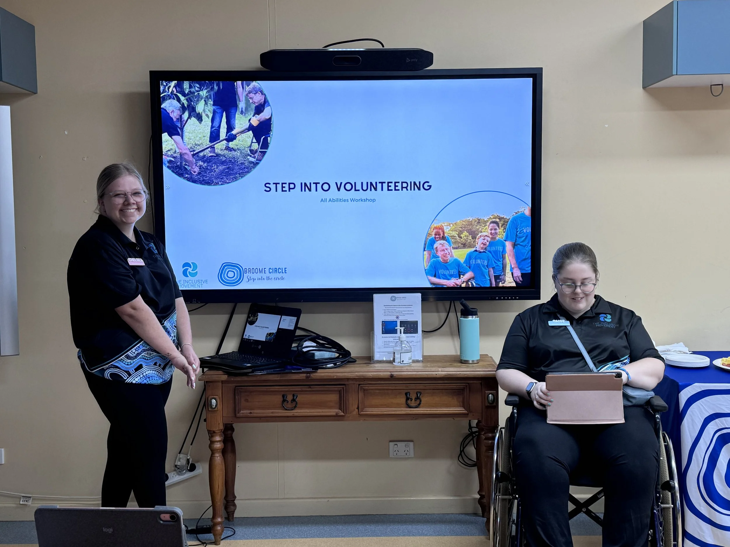 Two women, one standing and one sitting in a wheelchair, in a room with a large screen showing a presentation titled 'Step Into Volunteering' with photos of volunteers, a laptop, and various items on a wooden table.