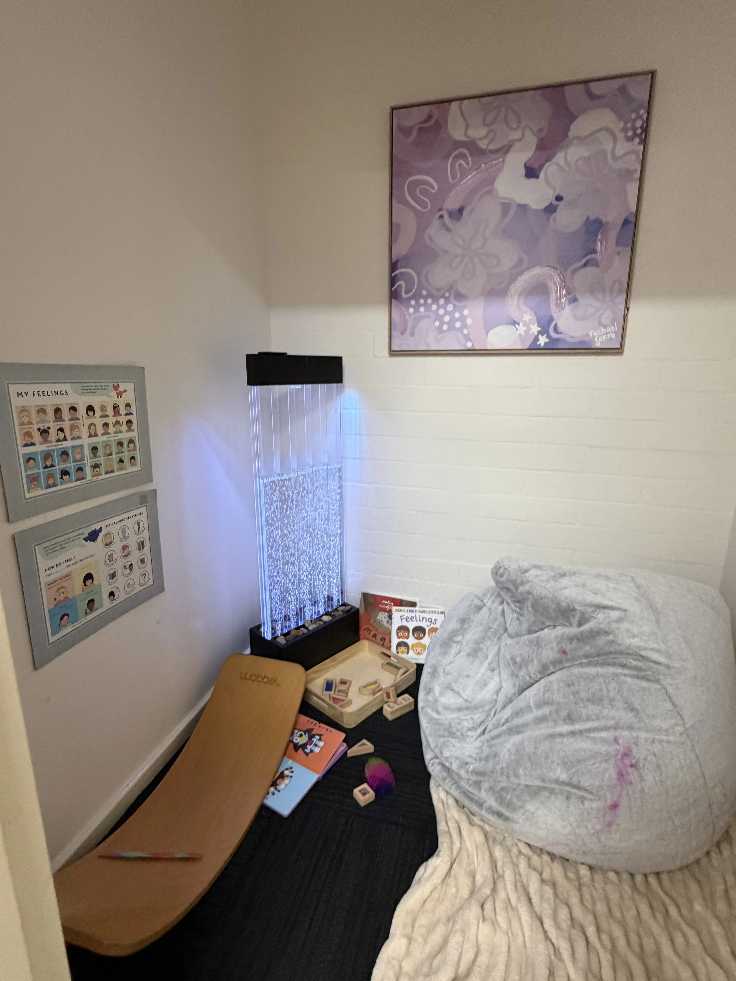 A cozy reading nook with a bean bag chair, a wooden lounge chair, and educational materials on the wall and floor. There's a small lighted water feature in the corner and a colorful abstract painting on the wall.