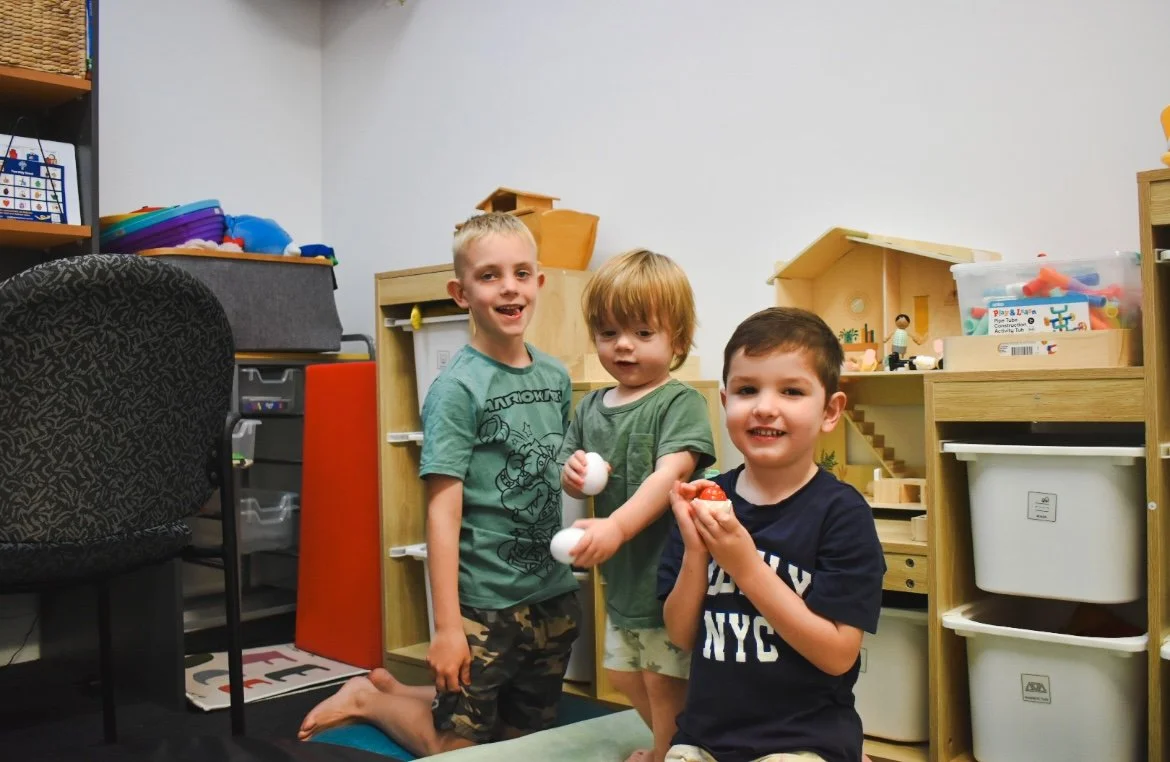 Three young boys playing inside a playroom with toys and furniture, smiling and holding eggs and a toy.