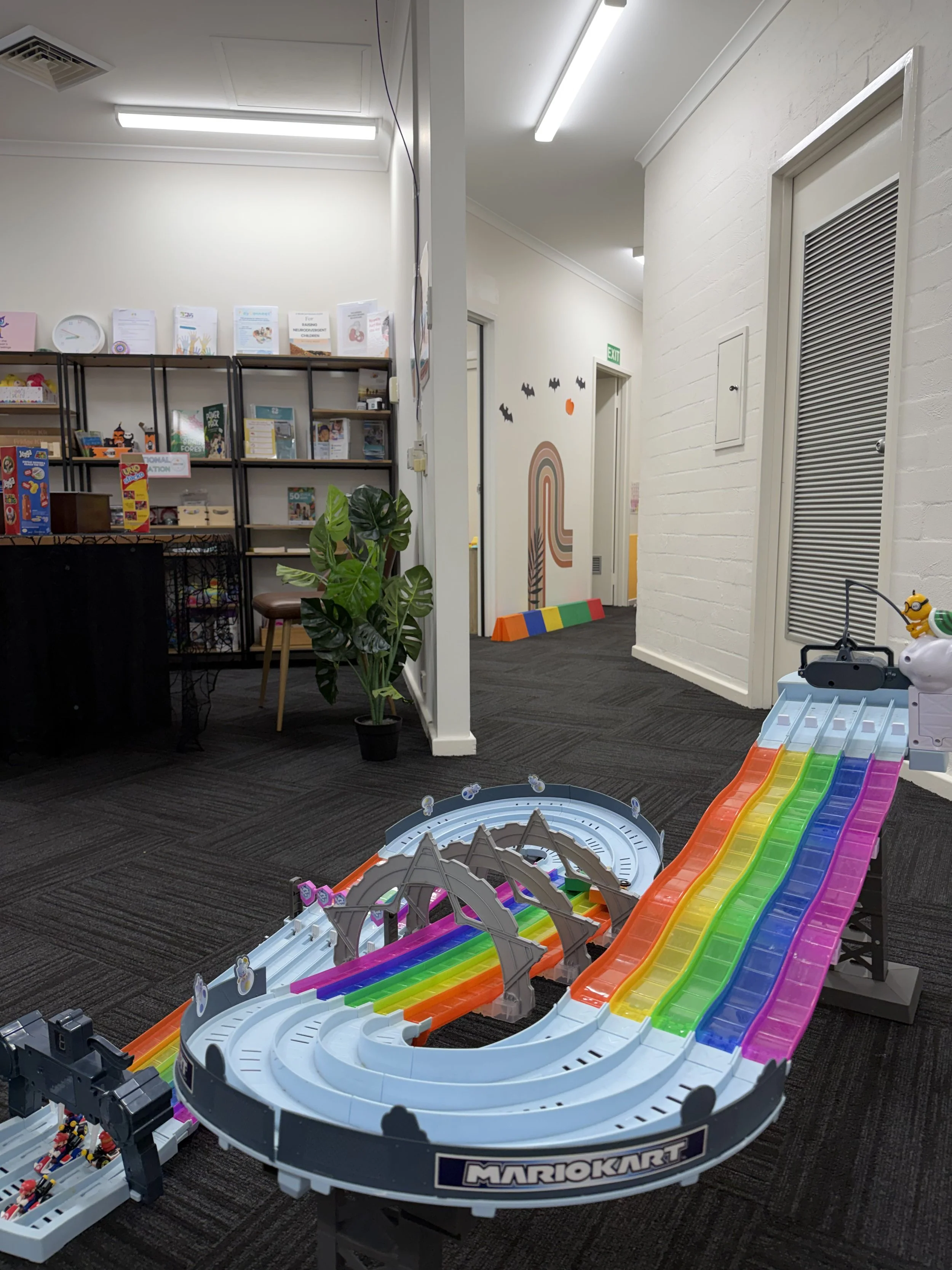Indoor play area with a colorful Mario Kart racing track toy on a dark carpeted floor, surrounded by white walls, a bookshelf, a square-shaped potted plant, and decorative wall art.