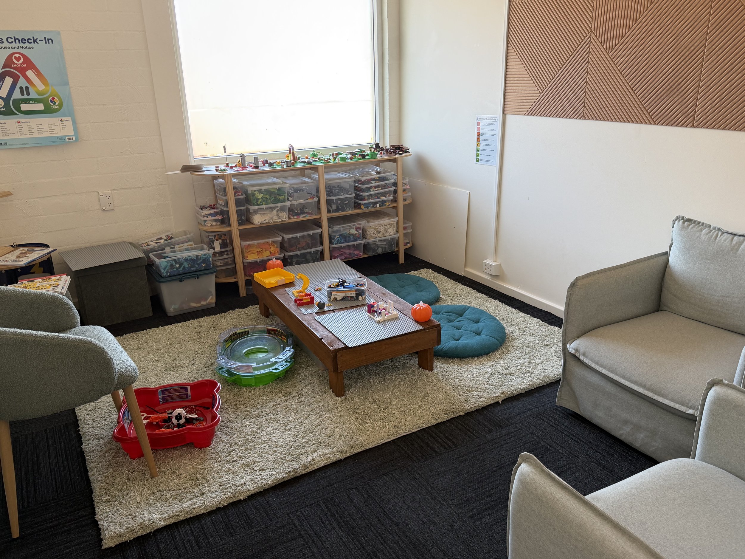 A cozy playroom with a beige rug, a low wooden table, blue floor cushions, a sofa, and shelves filled with Lego pieces and toys. A window lets in natural light.