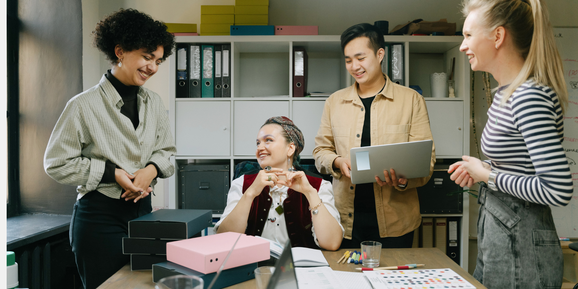 A diverse group of five young women working together in an office, standing and sitting around a table with documents, boxes, and office supplies, engaging in conversation and smiling.