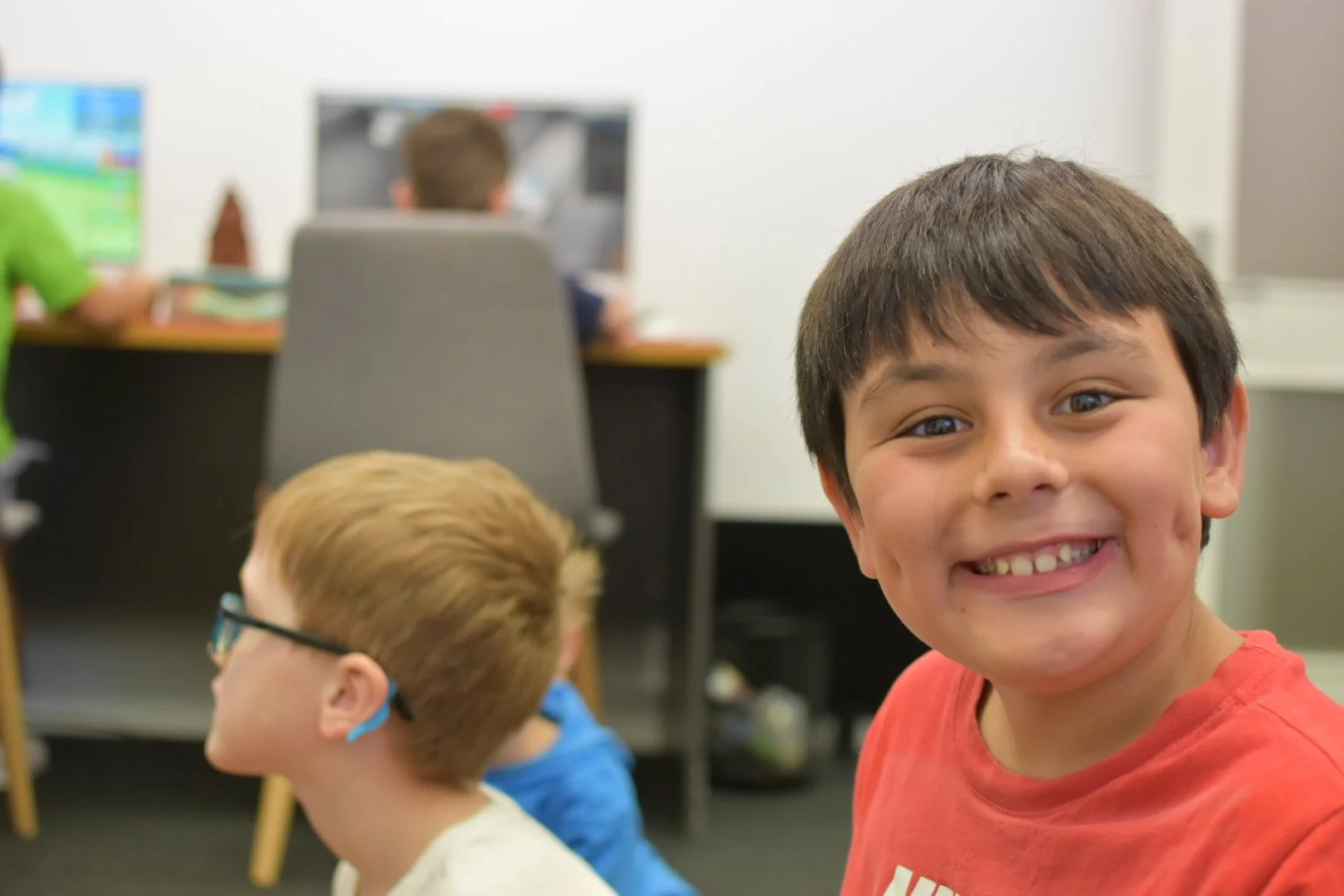 A happy young boy with dark hair and a red shirt smiling at the camera in a classroom with other children and computer monitors in the background.