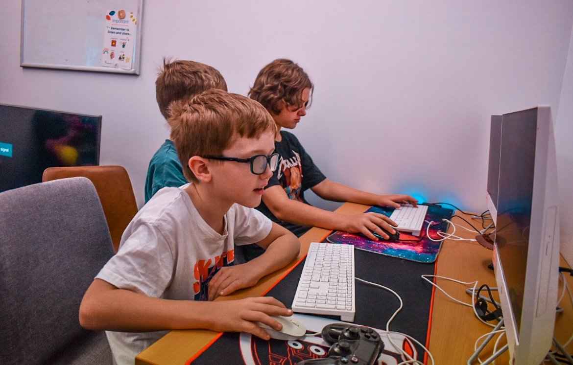 Three young boys playing computer games at a desk with monitors, keyboards, mice, and a game controller, in a room with a white wall and a poster.