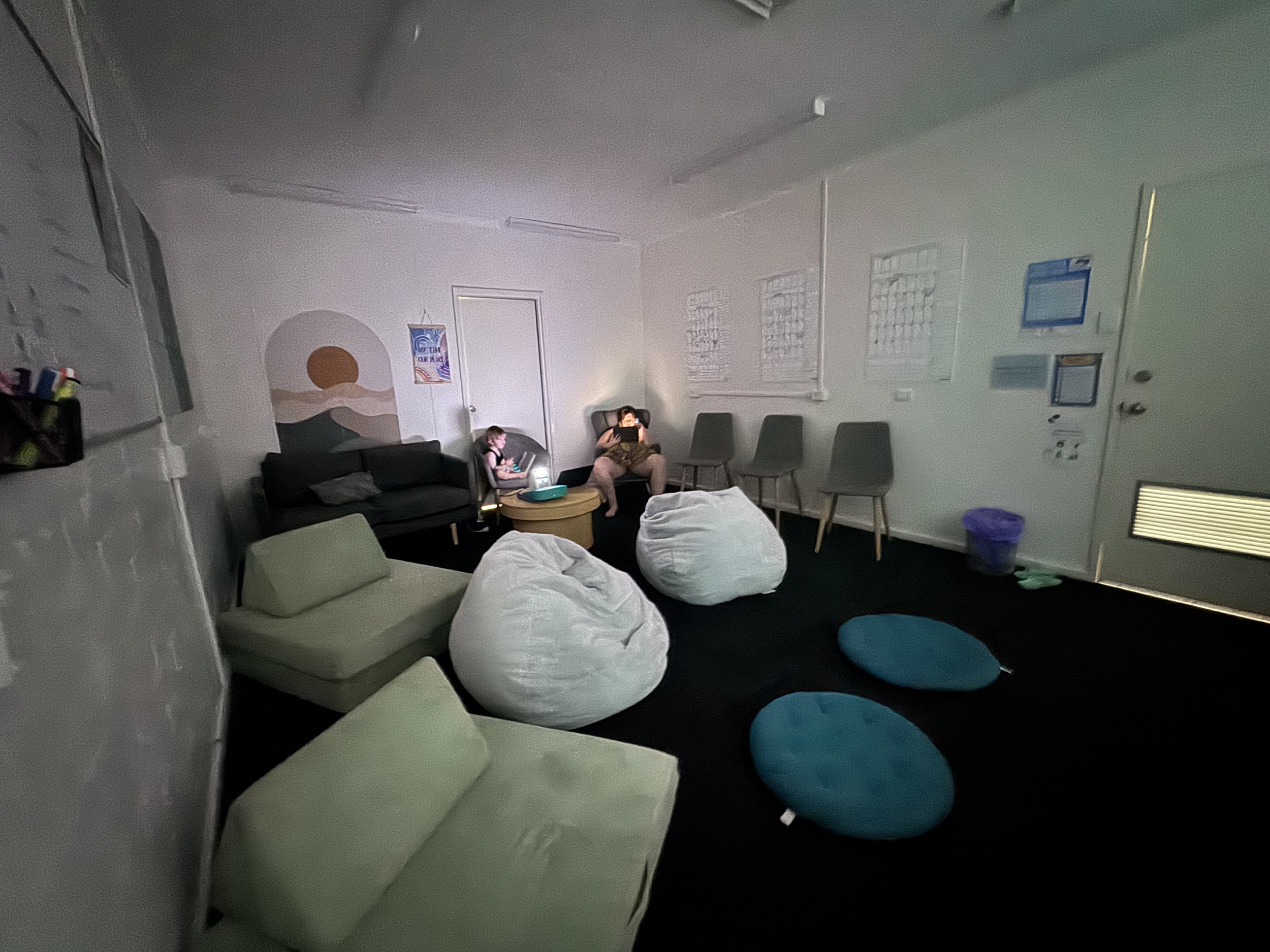 Indoor lounge with bean bag chairs, small sofas, and four blue floor cushions, two children sitting on a sofa and a bean bag, one child using a tablet, the room has white walls, windows with blinds, and some posters or notices on the wall.