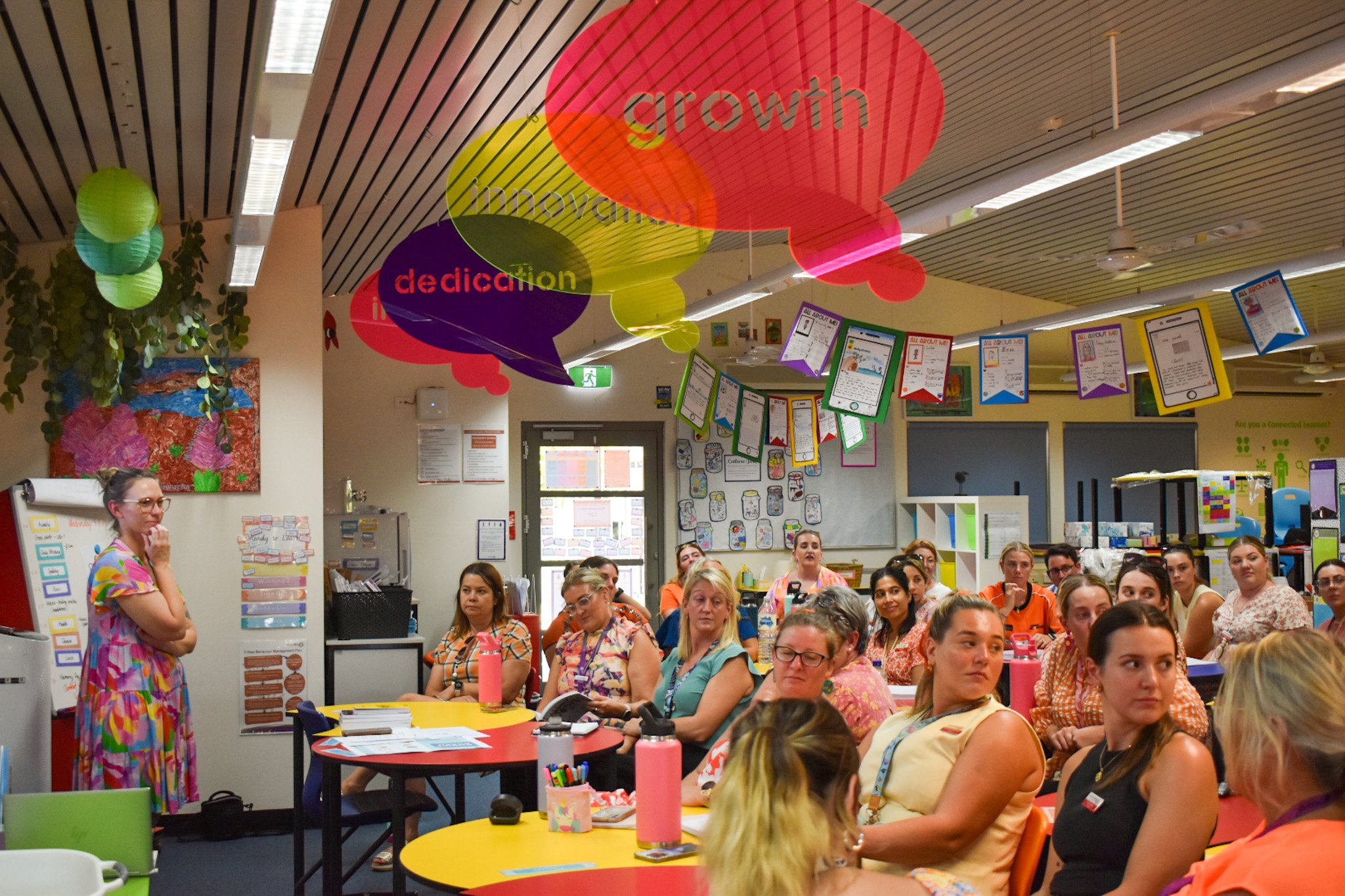 A teacher standing near the classroom wall and whiteboard, addressing a group of female adults seated at colorful tables in a classroom decorated with educational posters, hanging papers, and colorful lanterns from the ceiling.