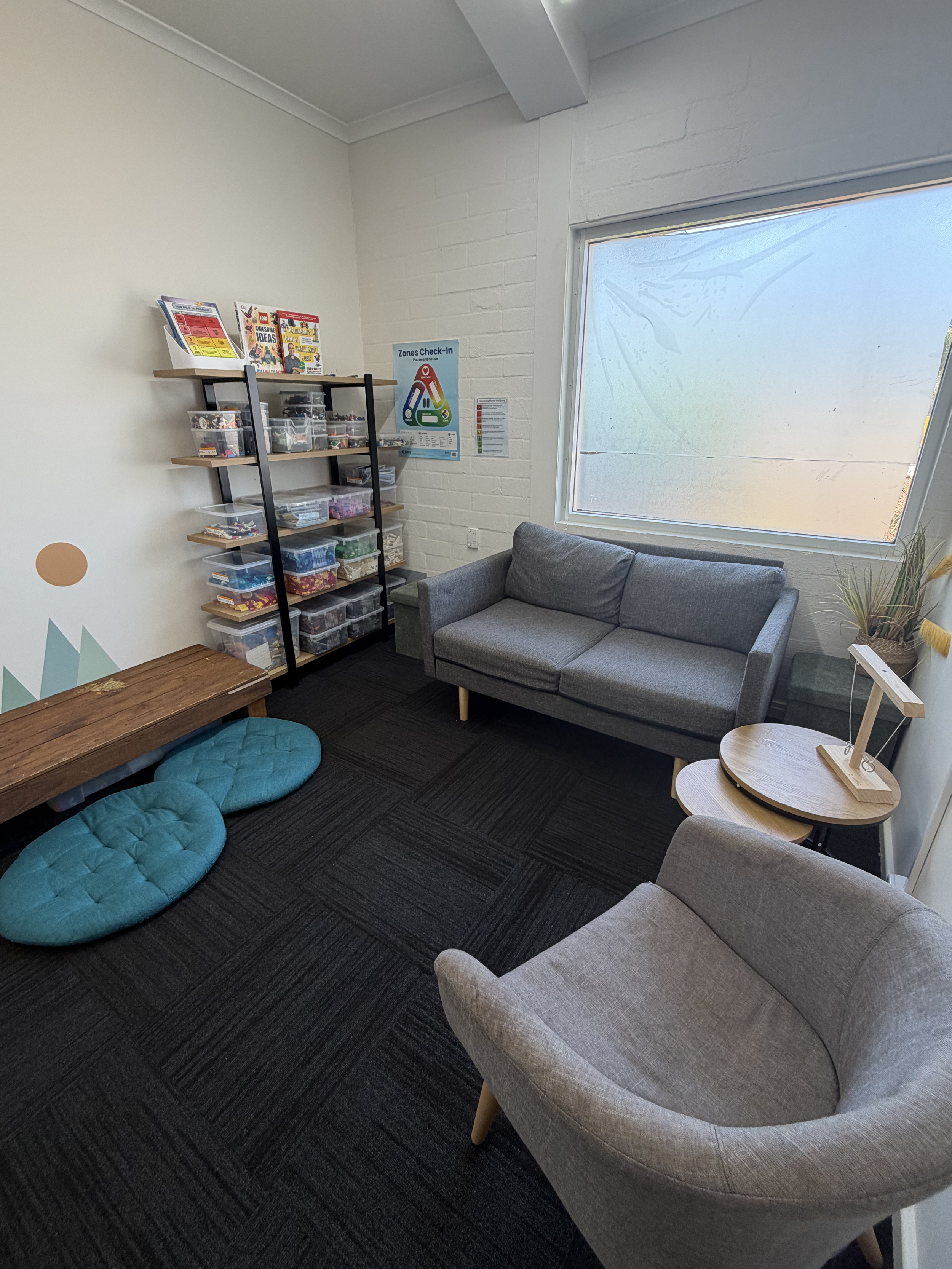 A small waiting room with a gray sofa, an armchair, two round wooden side tables, a wooden bench, teal floor cushions, a shelving unit with craft supplies, a window with frosted glass, and instructional posters on the wall.