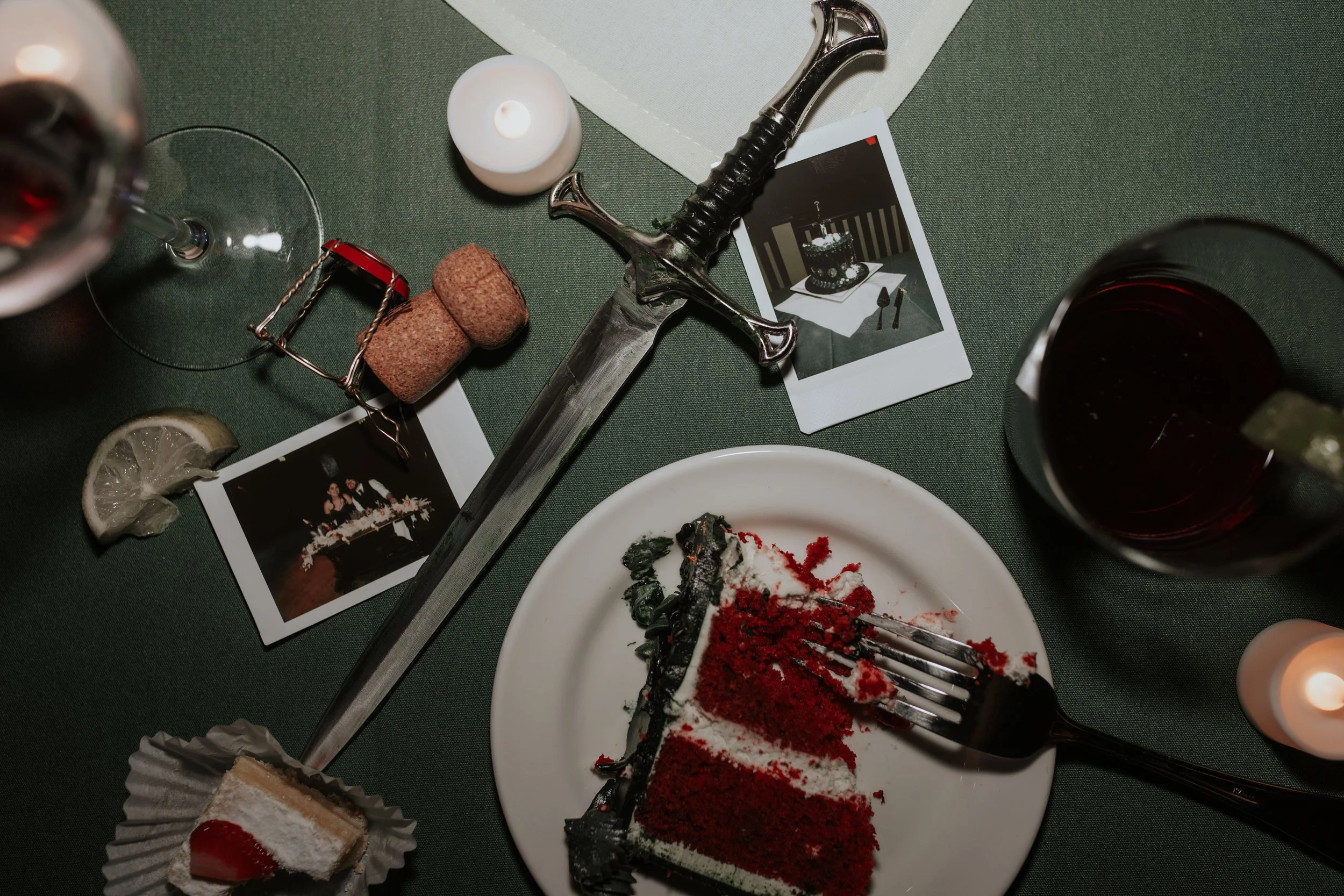 A partially eaten slice of red velvet cake on a white plate, a fork, wine glass, wine bottle, candle, photographs, and a sword on a green tablecloth, suggesting a dinner or celebration setting.