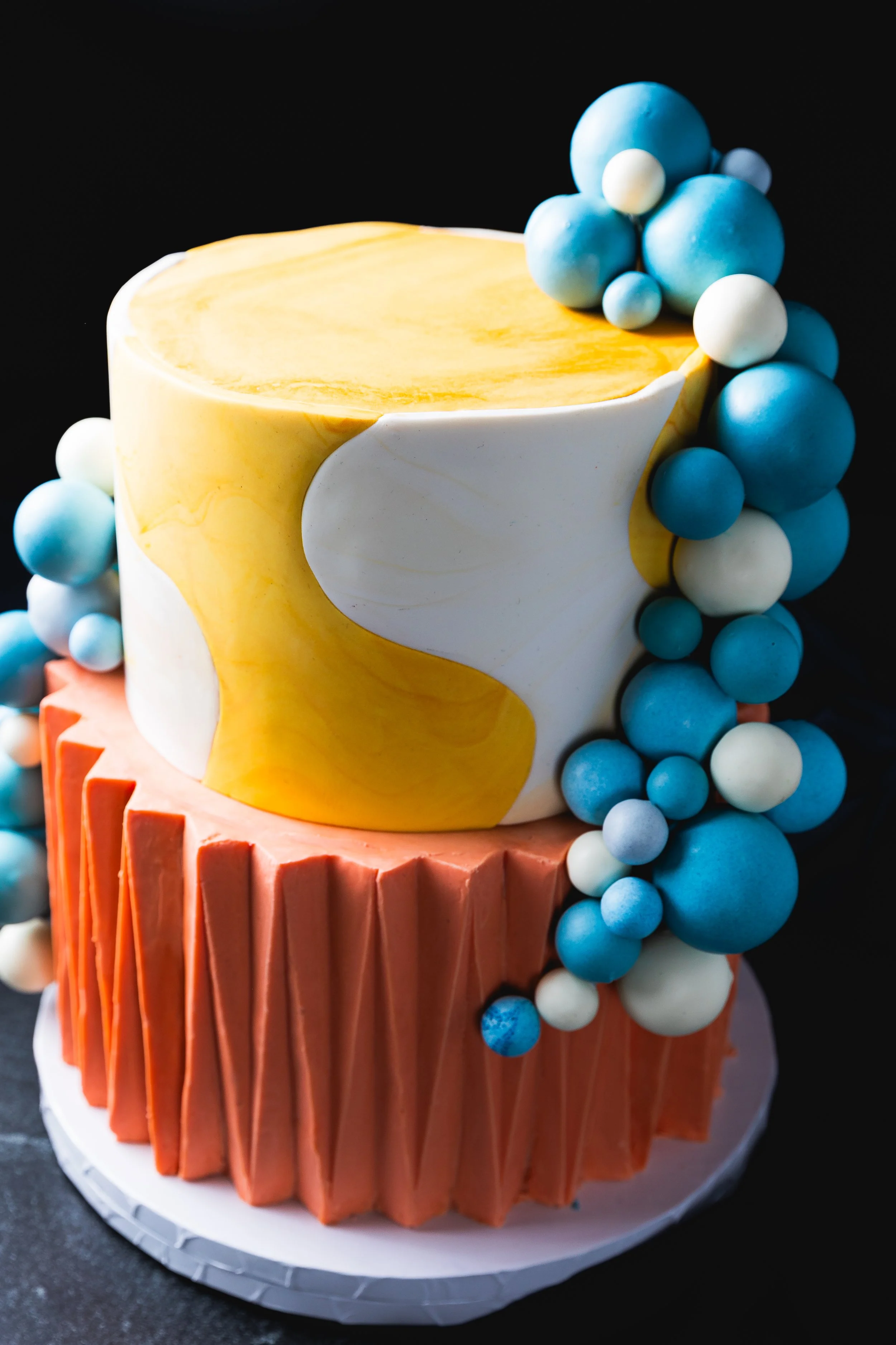 1960's themed cake with a marbled yellow and white top tier, orange origami style ganache base, and blue and white spherical accents on a black background.
