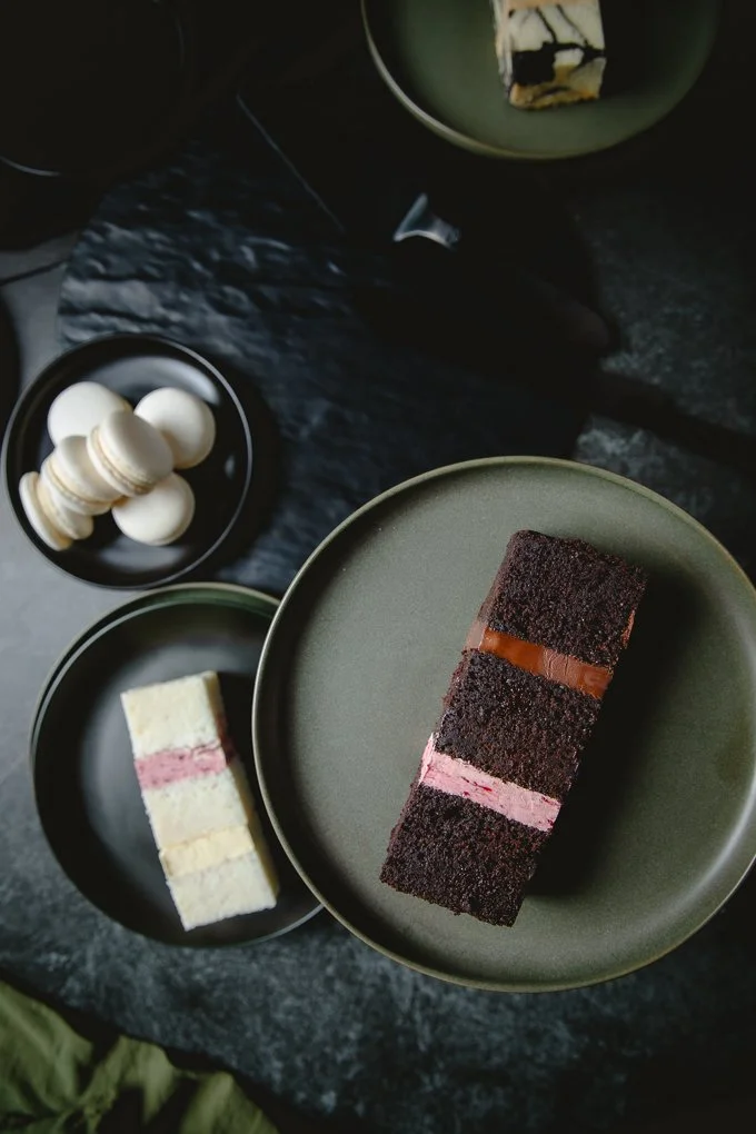 Assorted desserts on black and green plates, including a chocolate cake with pink filling, a layered vanilla and pink cake, and white macarons.