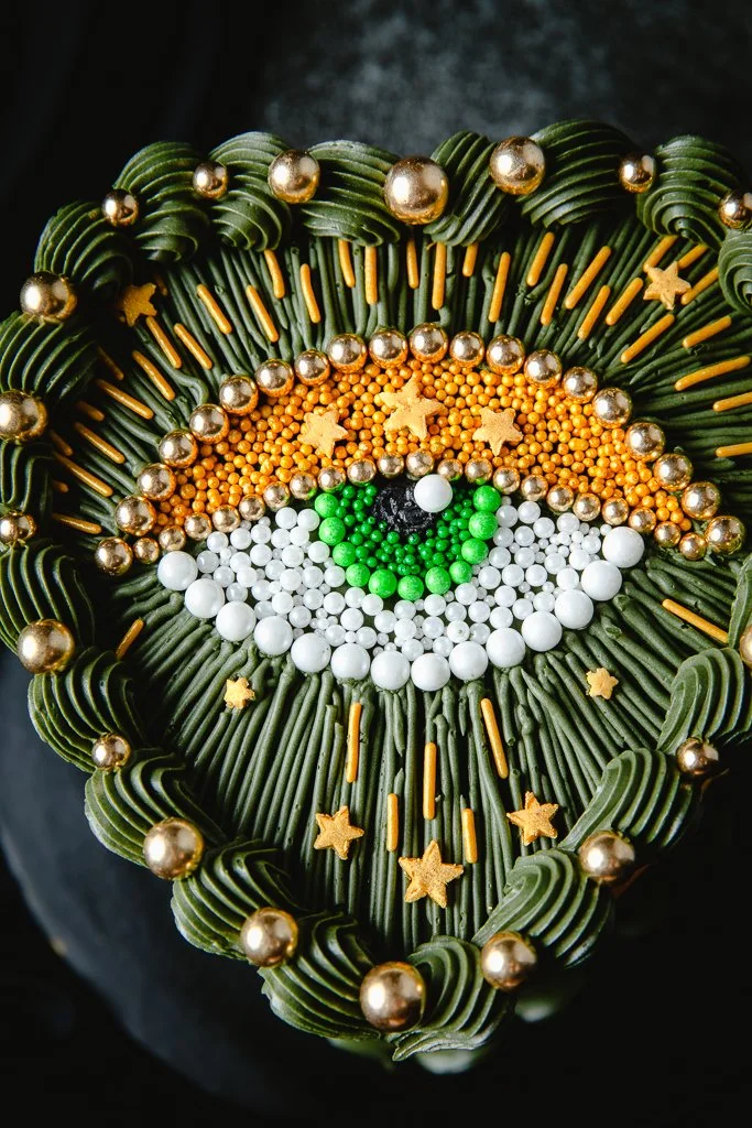 Vintage heart cake with evil eye motif, made of green, gold, white, and black candies and sprinkles, with star-shaped decorations.