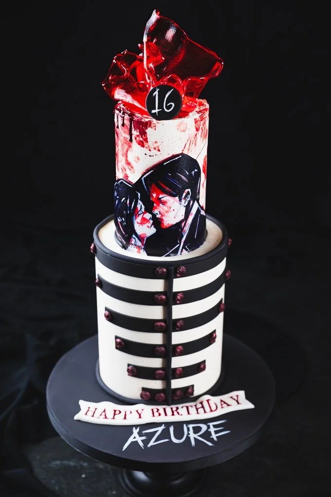 A birthday cake with a black and white striped cylindrical base, decorated with pink beads, and topped with a black and white depicting of two anime characters about to kiss, a black circle with the number 16, a glass filled with red liquid, ice, and an illustrated cutout of two anime characters in an intimate pose. The cake has a white banner with red letters saying "HAPPY BIRTHDAY" and the name "AZURE" written below.