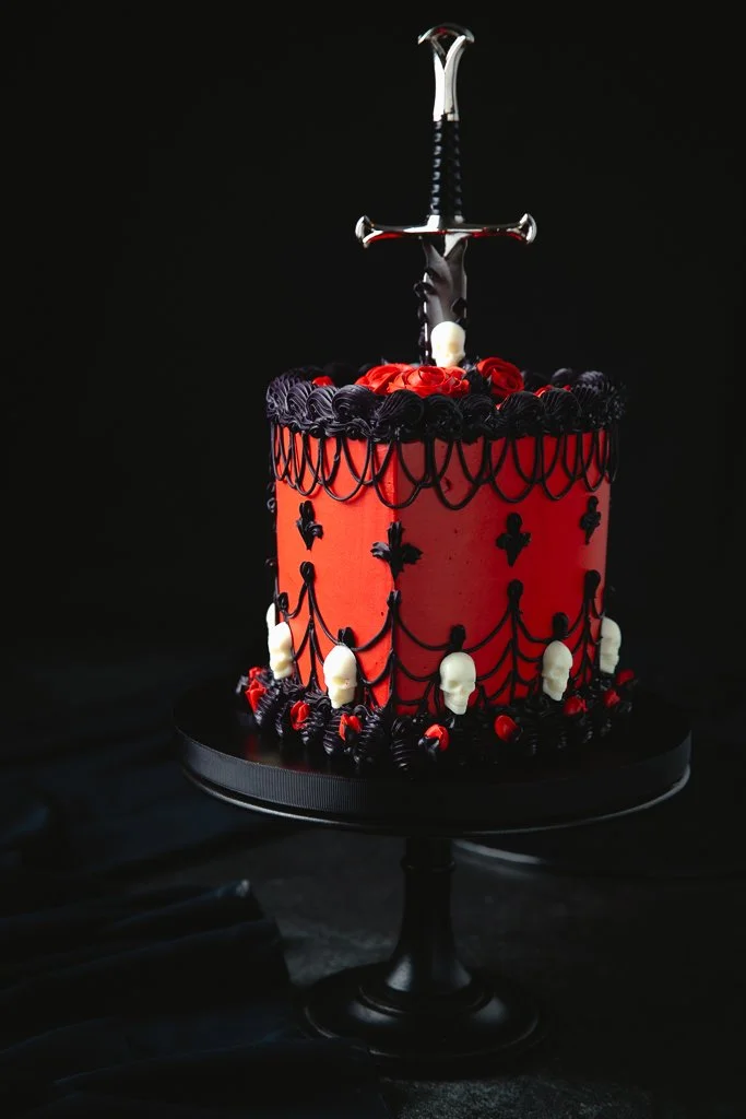 A tall, red and black cake decorated with skull and crossbones motifs, black and red piping, topped with a small sword with a black and silver handle.