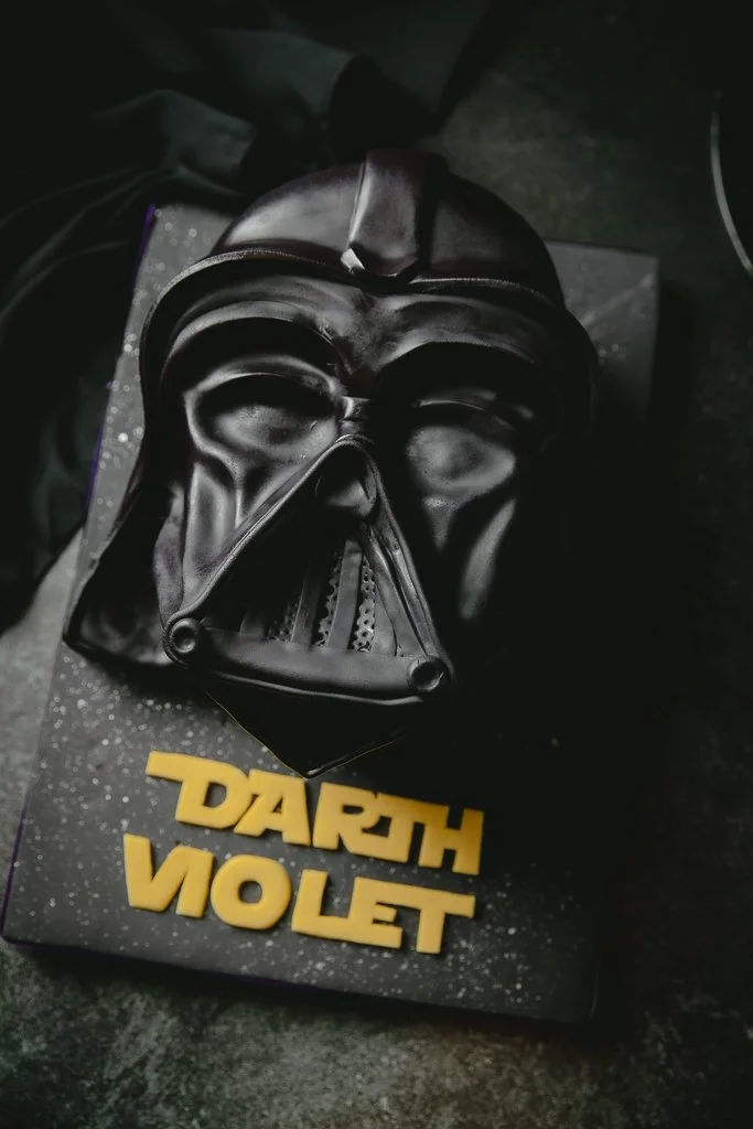 A Darth Vader mask replica resting on a box with "Darth Vader" written in yellow letters, placed on a dark surface.