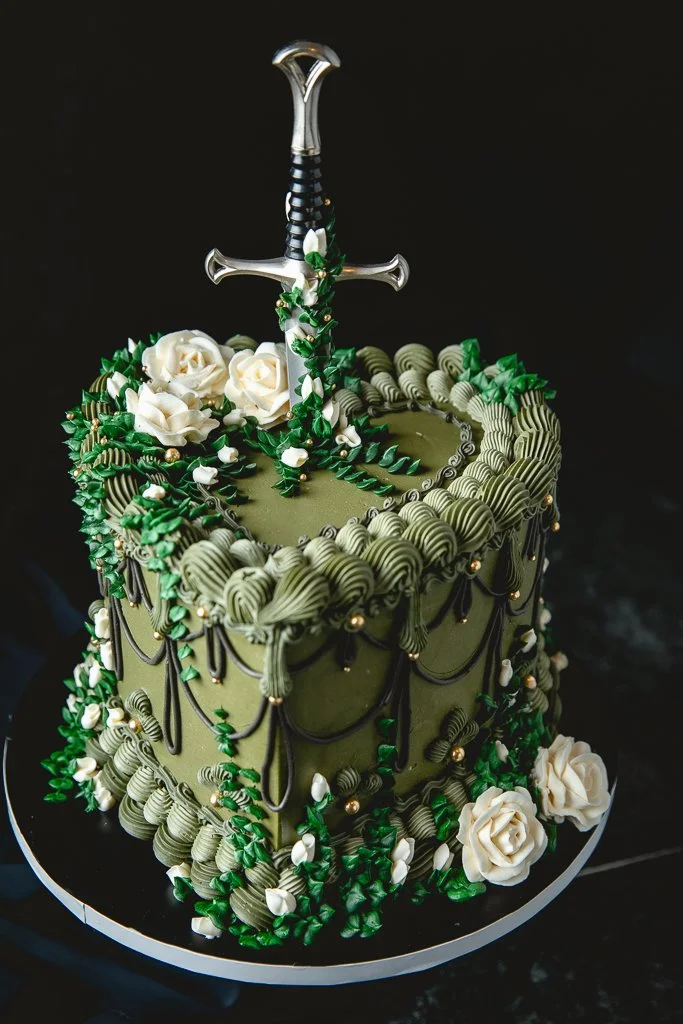 Vintage olive green cake with white buttercream roses, trailing moss, and a dagger cake topper.