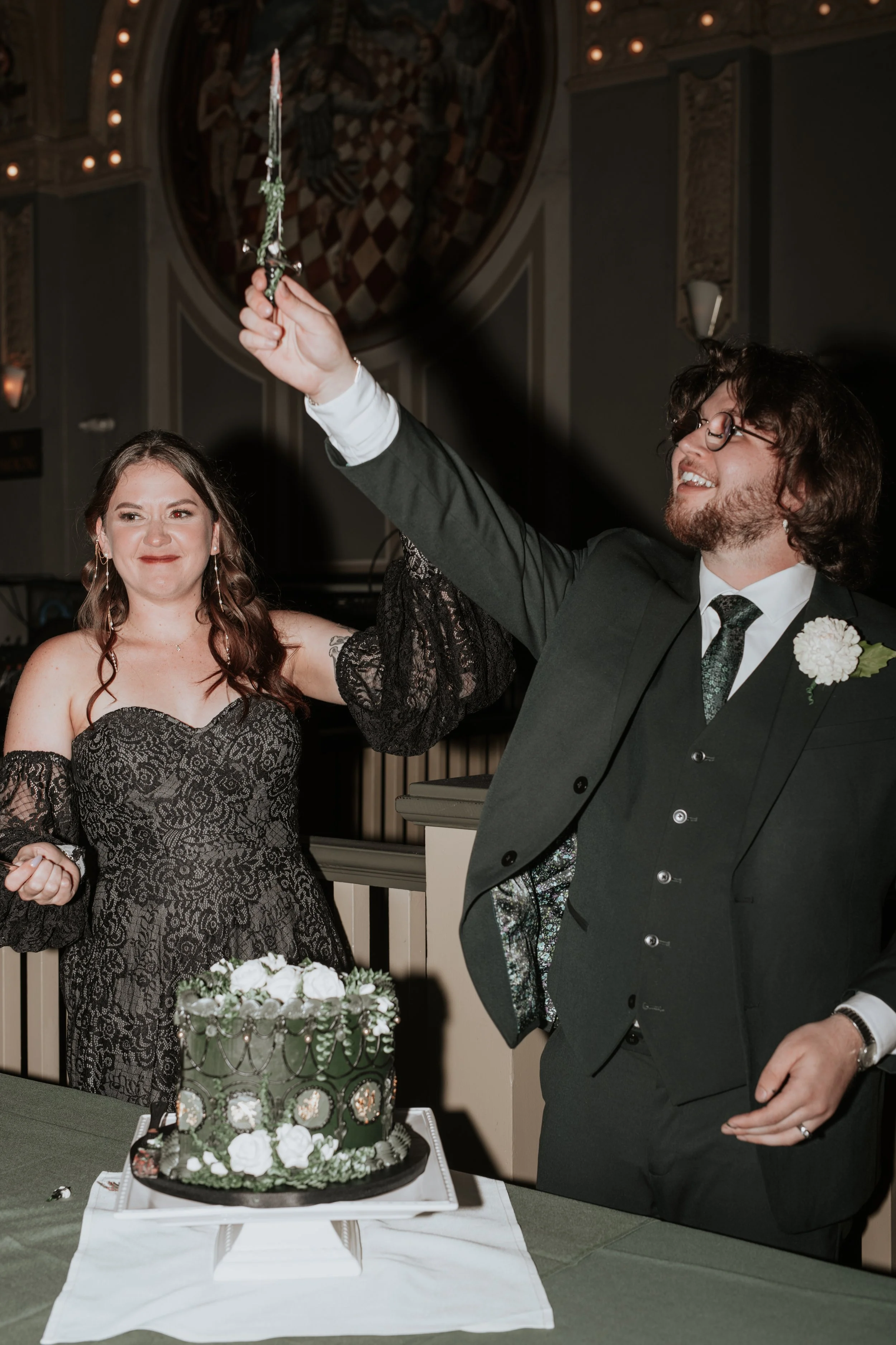 A groom holds a dagger aloft above a vintage cutting cake. His bride stands next to him in a black lace wedding dress, smiling.
